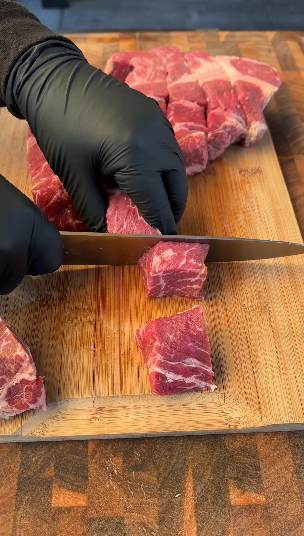Chuck roast being cut into 1-inch cubes on a bamboo cutting board with black gloved hands for honey BBQ burnt ends