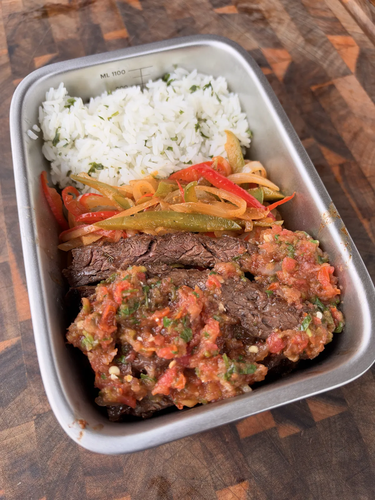 Meal prep steel tray with cilantro lime rice, charred bell peppers and onions, sliced skirt steak topped with homemade salsa