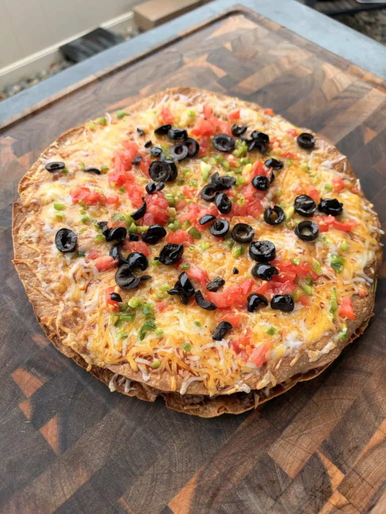 Homemade Taco Bell copycat Mexican pizza with melted Colby Jack cheese, diced tomatoes, sliced black olives, and jalapeños on a wood cutting board