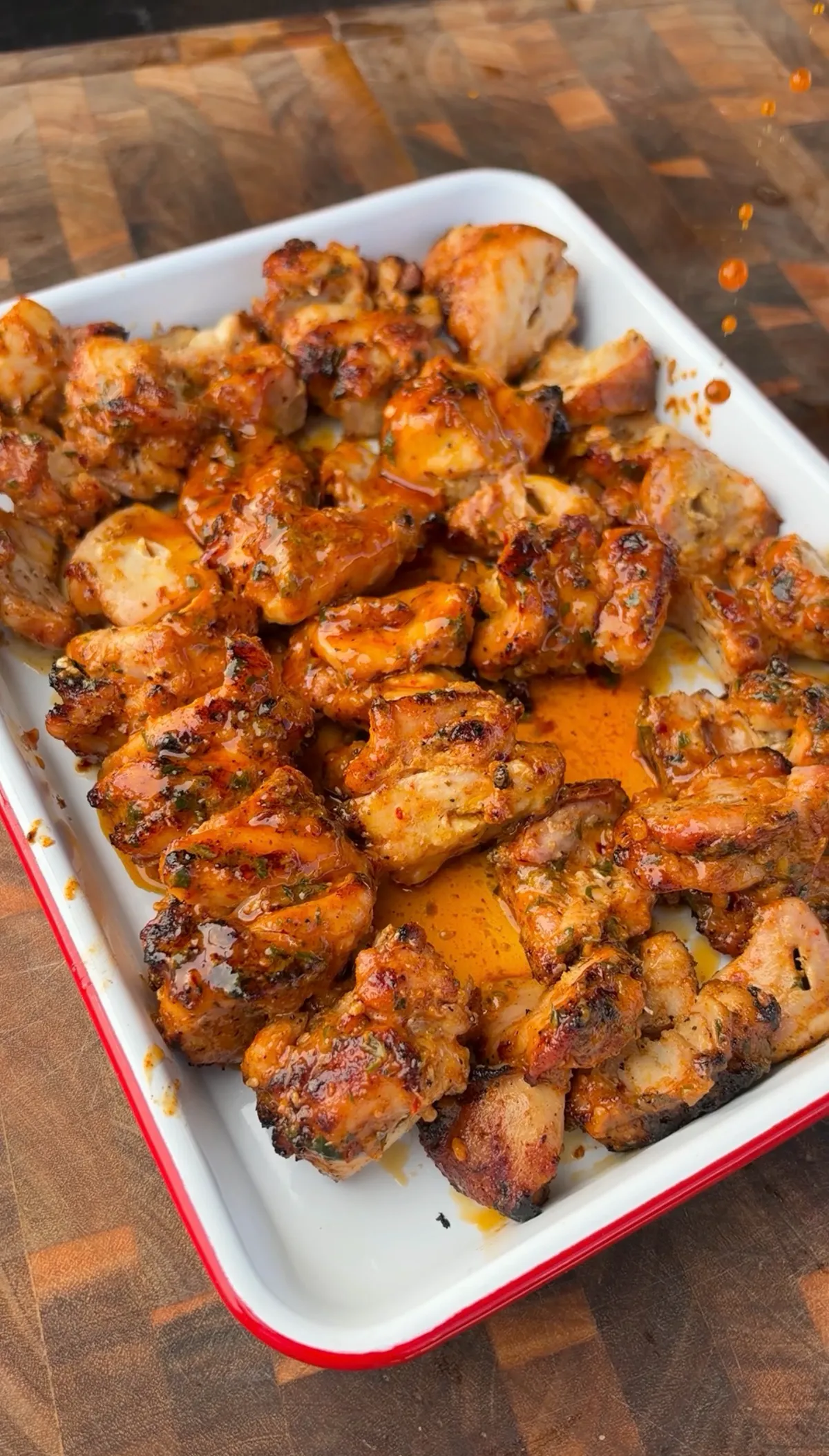 Grilled chicken thigh pieces coated in glossy cowboy butter sauce served in a white enamel tray on a wood cutting board
