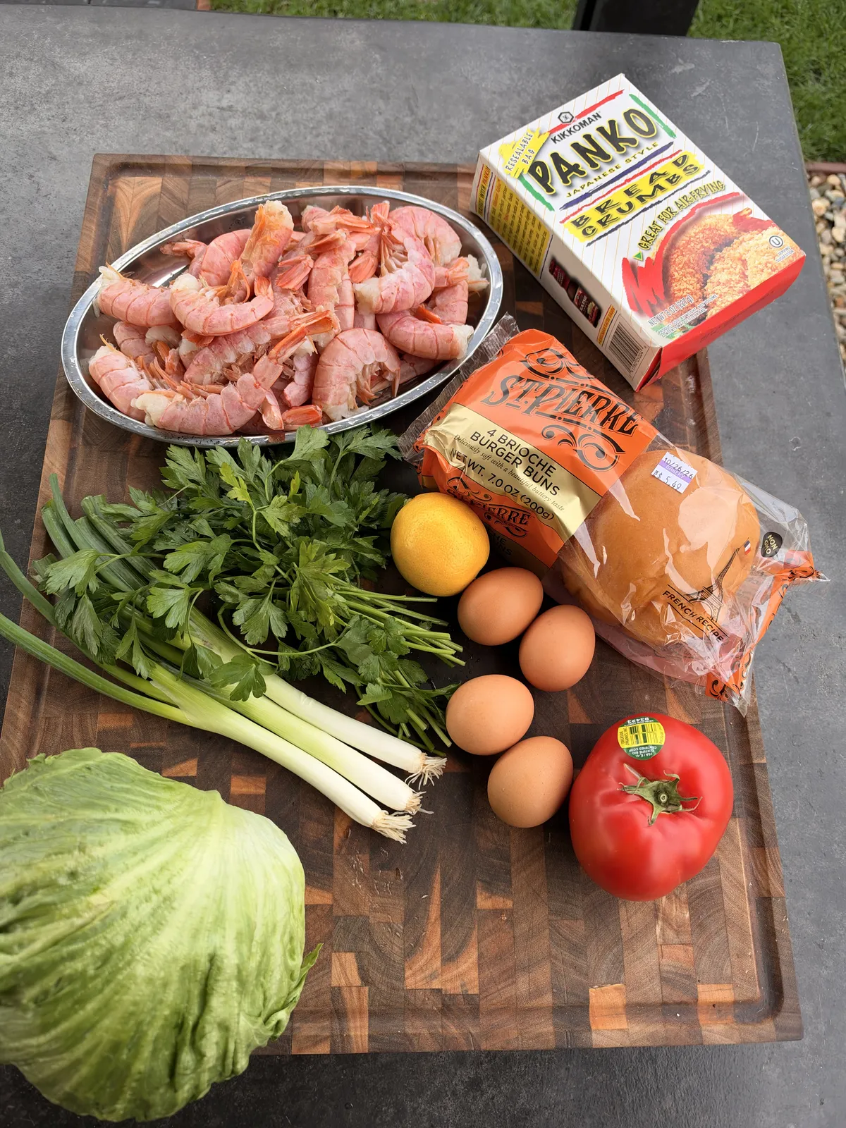 Raw Argentine shrimp, panko breadcrumbs, eggs, brioche buns, parsley, green onions, lemon, lettuce, and tomato on a wooden cutting board