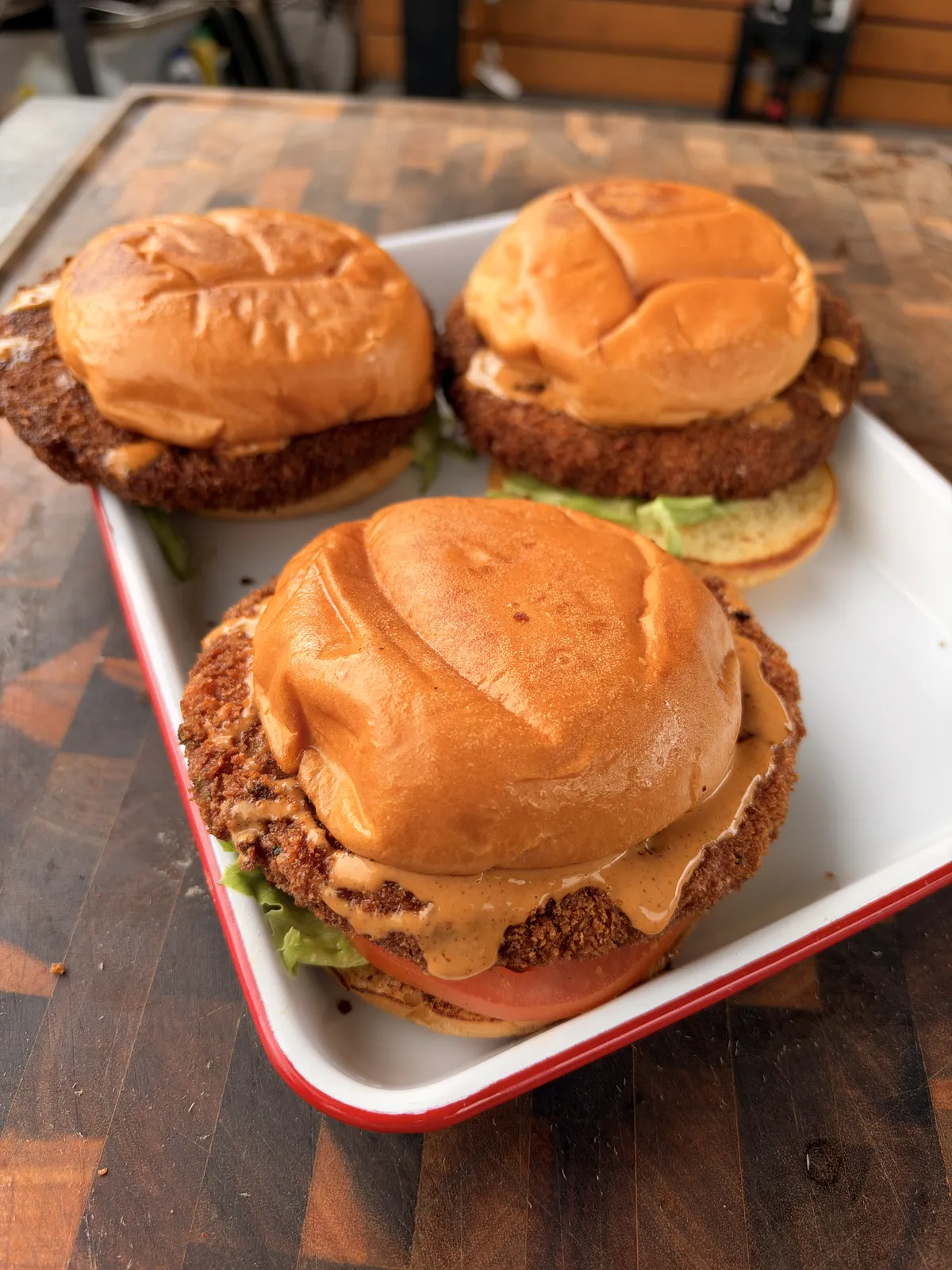 Three crispy fried shrimp burgers on brioche buns with chipotle mayo, lettuce, and sliced tomato on a white tray