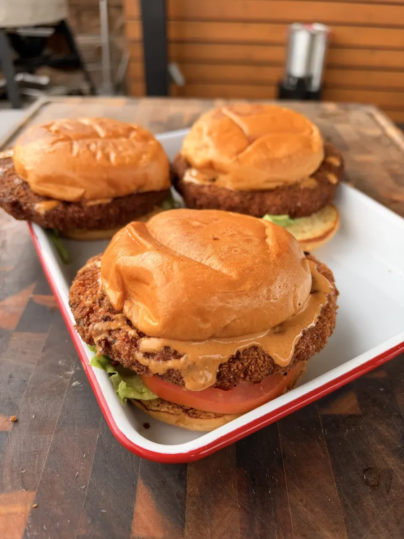 Crispy fried shrimp burgers on toasted brioche buns with chipotle mayo, lettuce, and sliced tomato in a white enamel tray