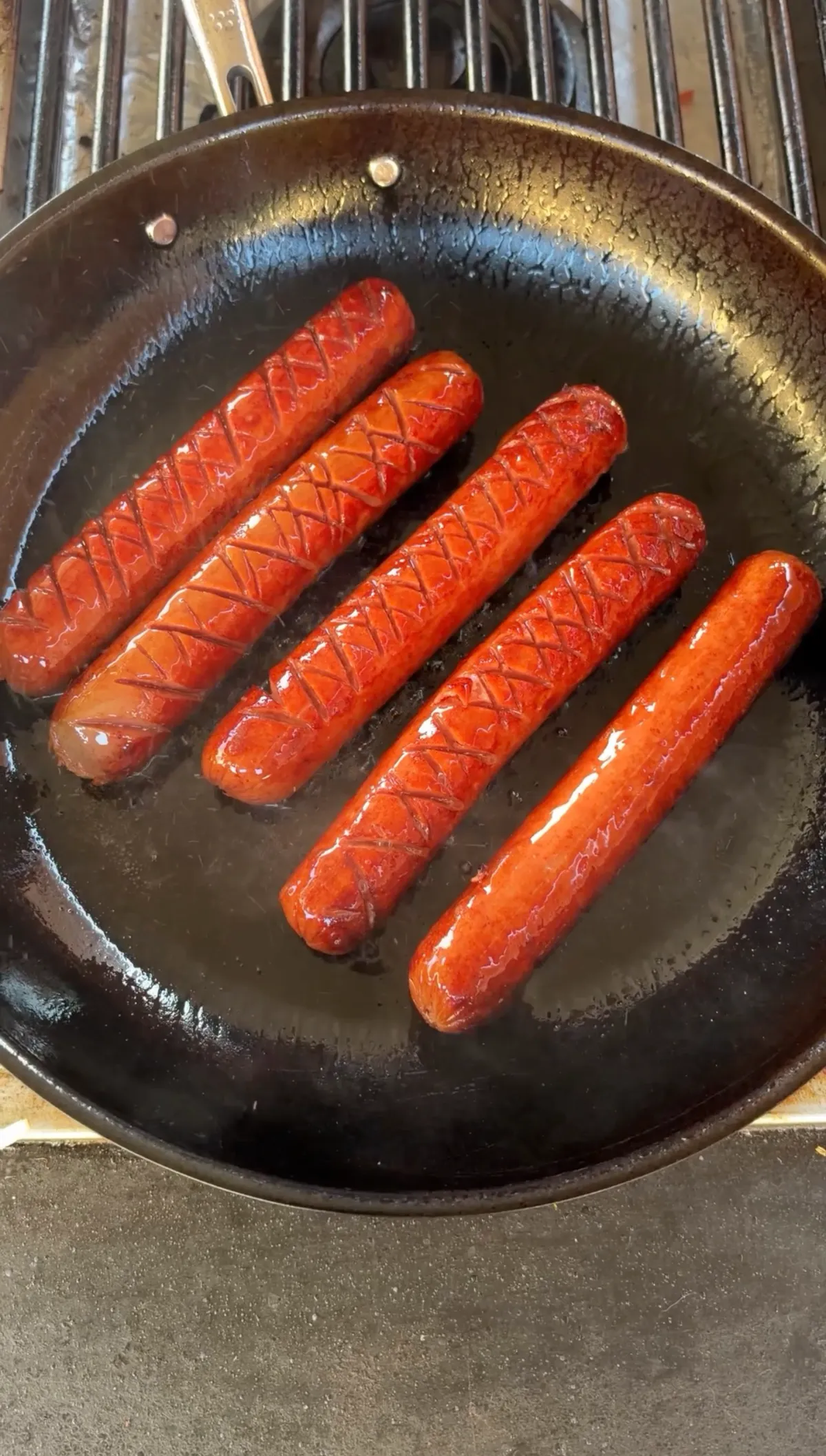 Five crosshatch-scored hot dogs frying in a dark skillet until browned and cuts begin to open