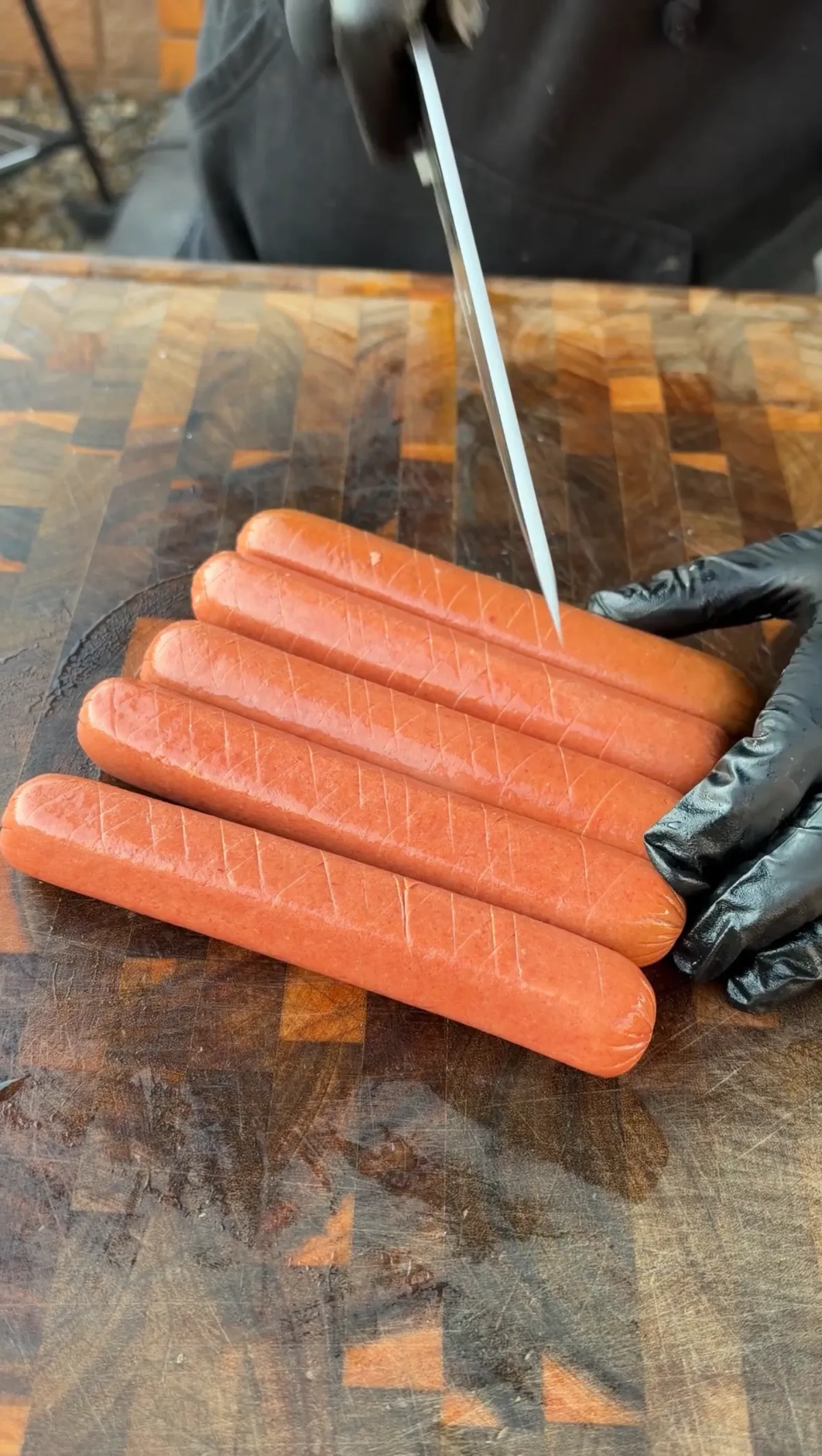 Five raw hot dogs with crosshatch scoring pattern being cut on a wooden cutting board with black gloved hands