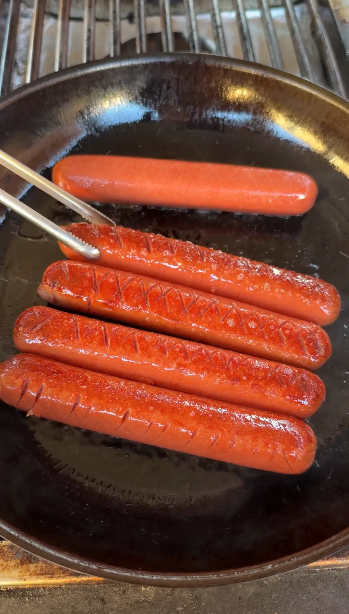 Five hot dogs with crosshatch score patterns frying in a cast iron skillet, held by tongs
