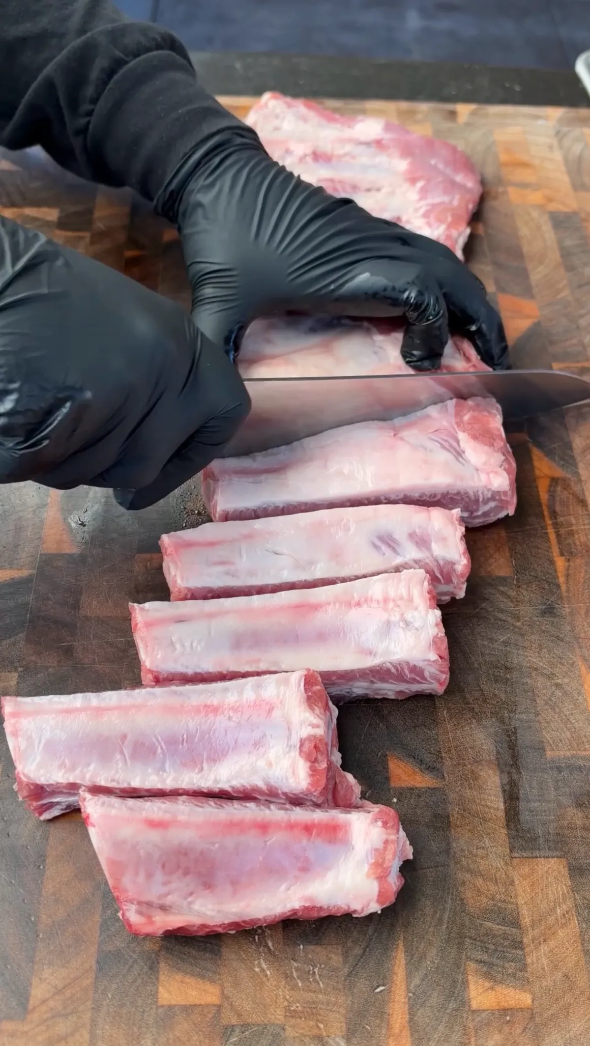 Gloved hands using a knife to cut St. Louis-style pork ribs individually between the bones on a wooden cutting board