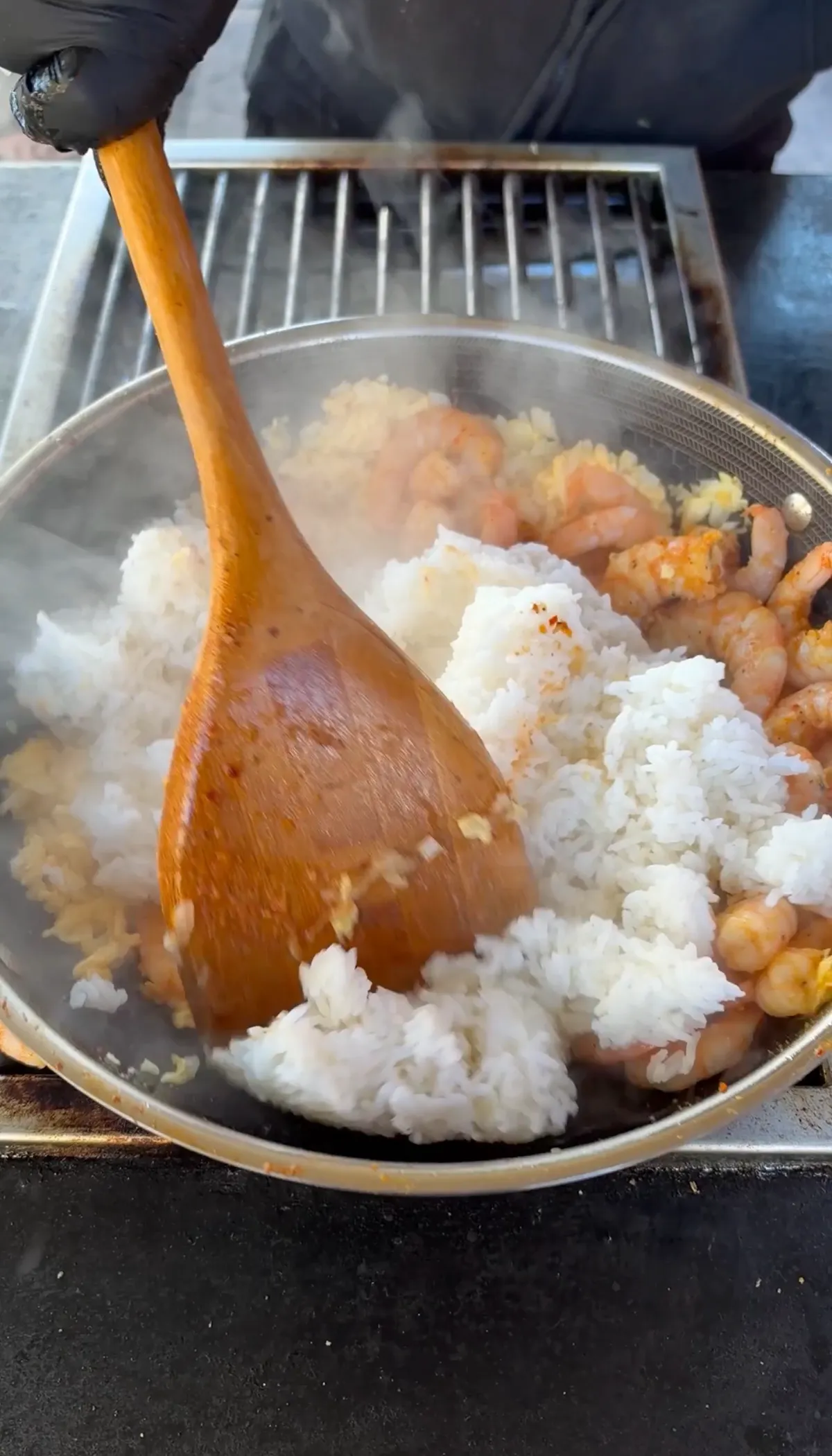 Day-old white rice and cooked shrimp being tossed together in a stainless wok with a wooden spoon