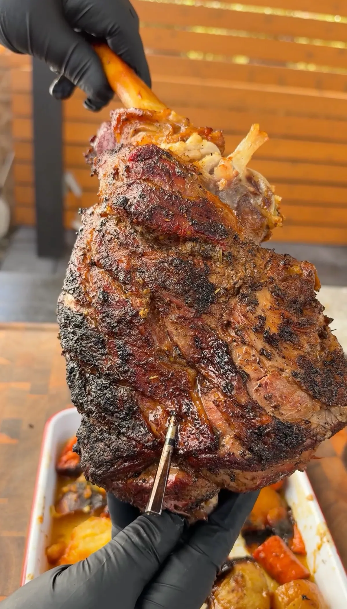 Hands in black gloves lifting a fall-apart tender braised leg of lamb with MEATER thermometer inserted over roasting tray