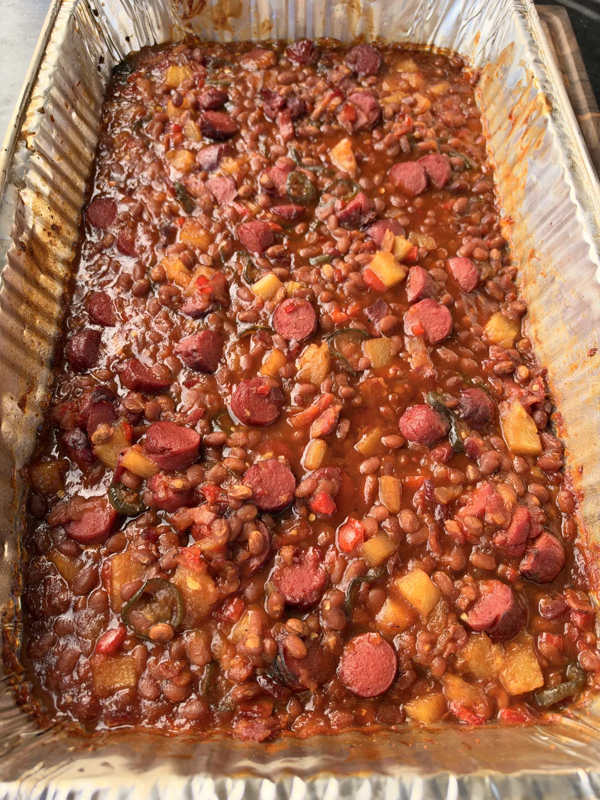 Smoked BBQ baked beans with sliced hot dog rounds, jalapeños, and pineapple in a foil pan