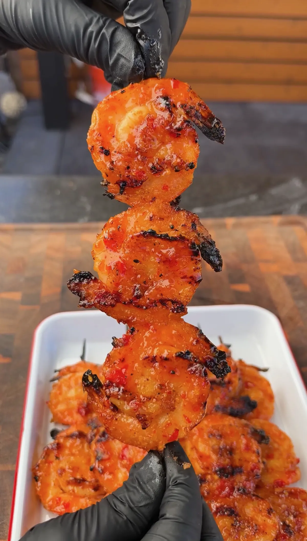 Gloved hands holding a skewer of caramelized firecracker-glazed grilled shrimp in yin-yang shape over a white dish