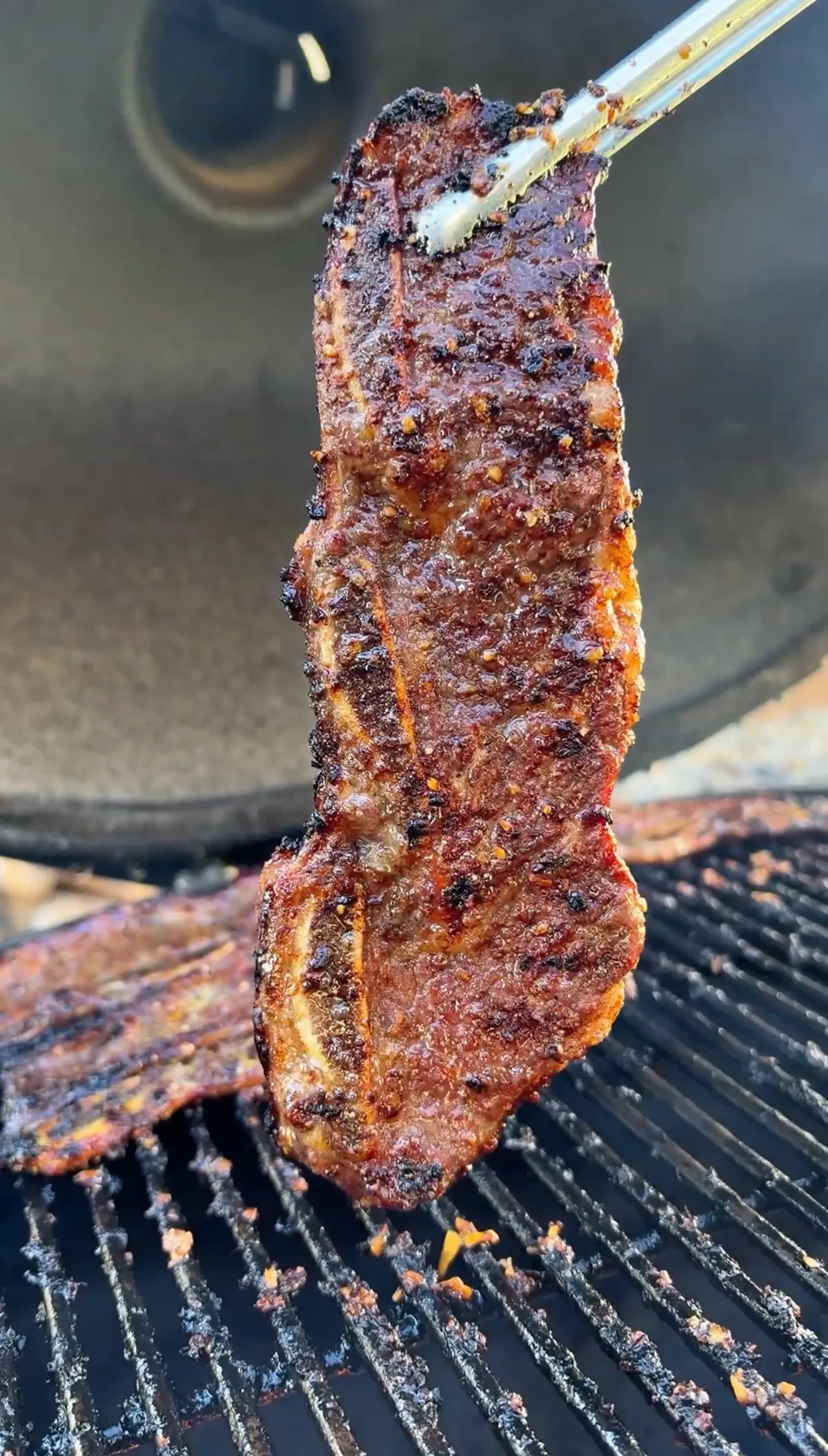 Tongs lifting a flanken-cut beef short rib with deep caramelized char and kalbi marinade crust over hot grill grates