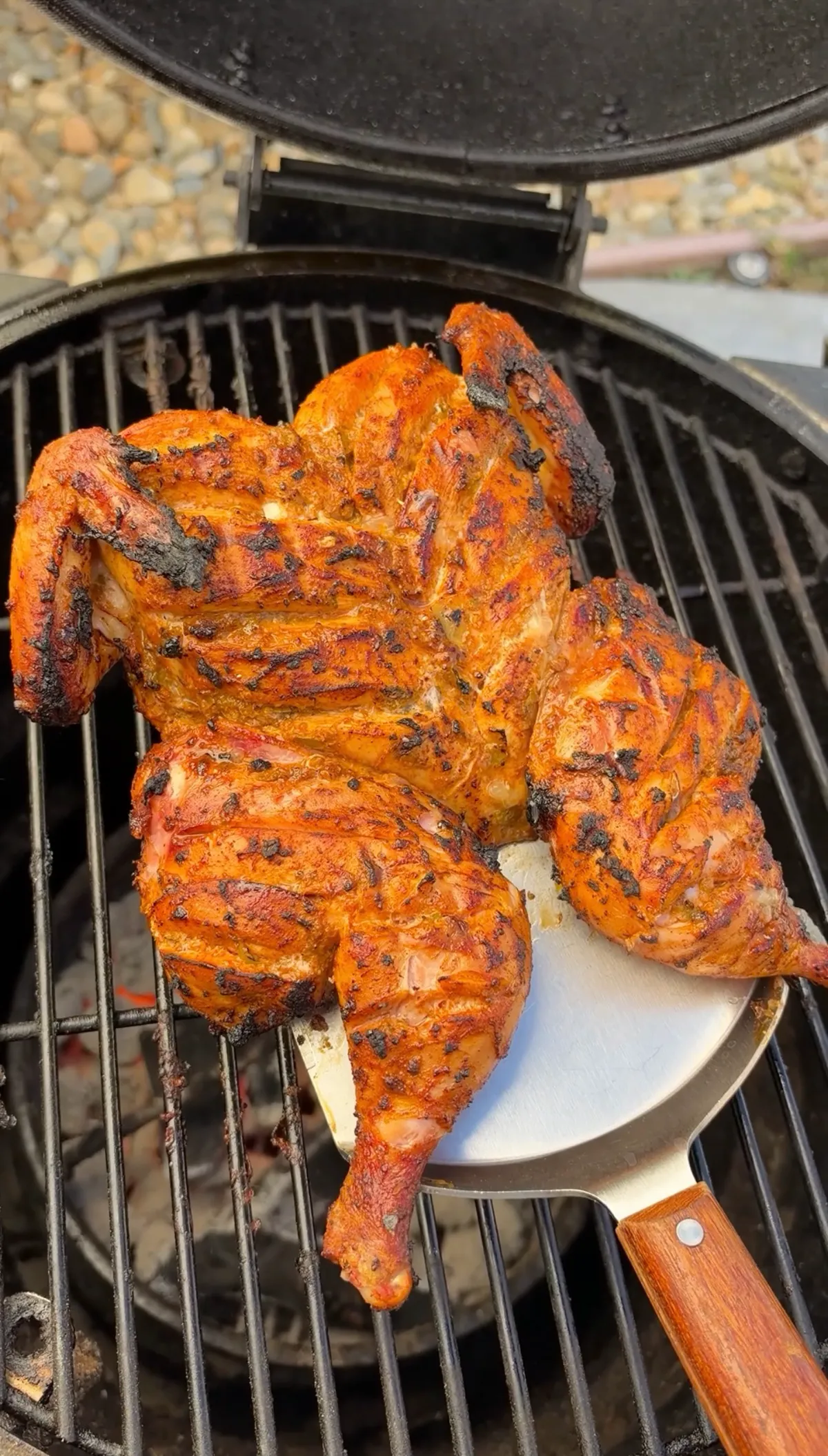 Spatchcocked green chile lime chicken being lifted with a spatula on a charcoal grill, showing charred scored skin