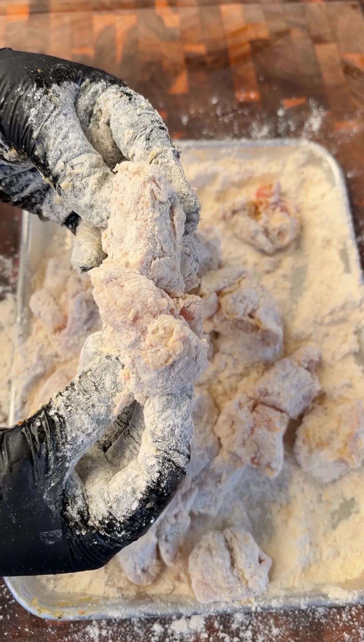 Black-gloved hand holding flour-coated chicken thigh nuggets over a tray of all-purpose flour before frying