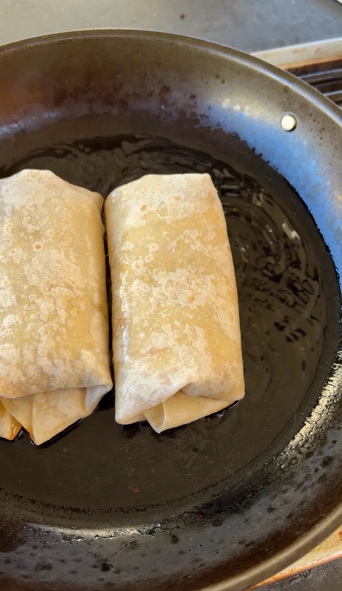 Two folded flour tortilla cheeseburger burritos frying in oil in a dark shallow pan until golden brown