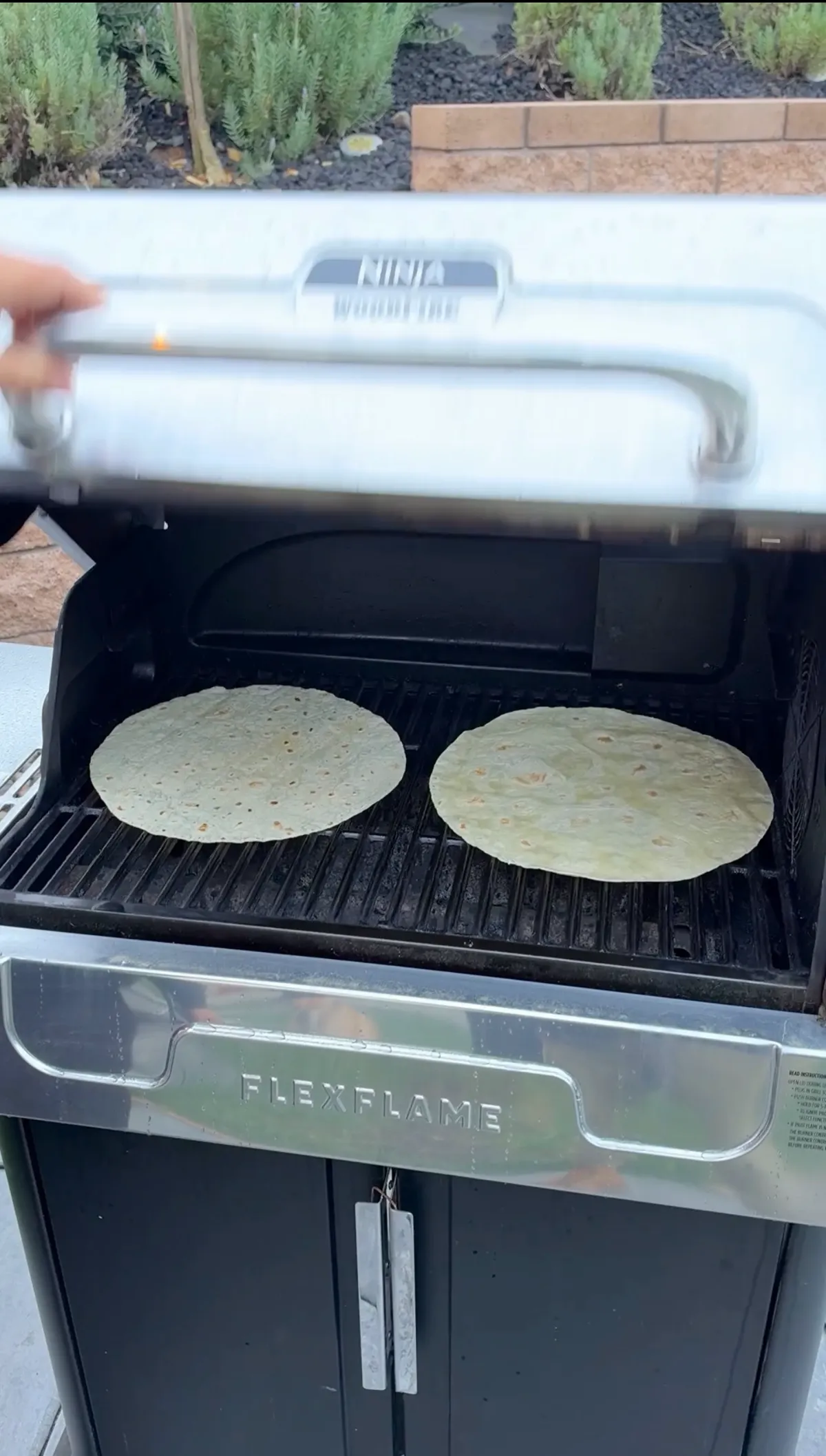 Two large burrito-size flour tortillas crisping on a Ninja Woodfire Flexflame grill grates outdoors