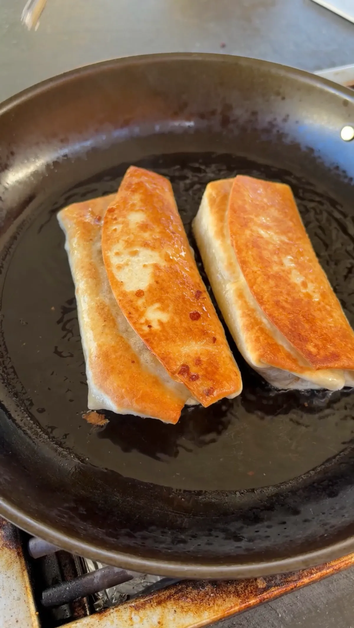 Two pan-fried ground beef burritos browning in oil in a skillet, golden brown on all sides
