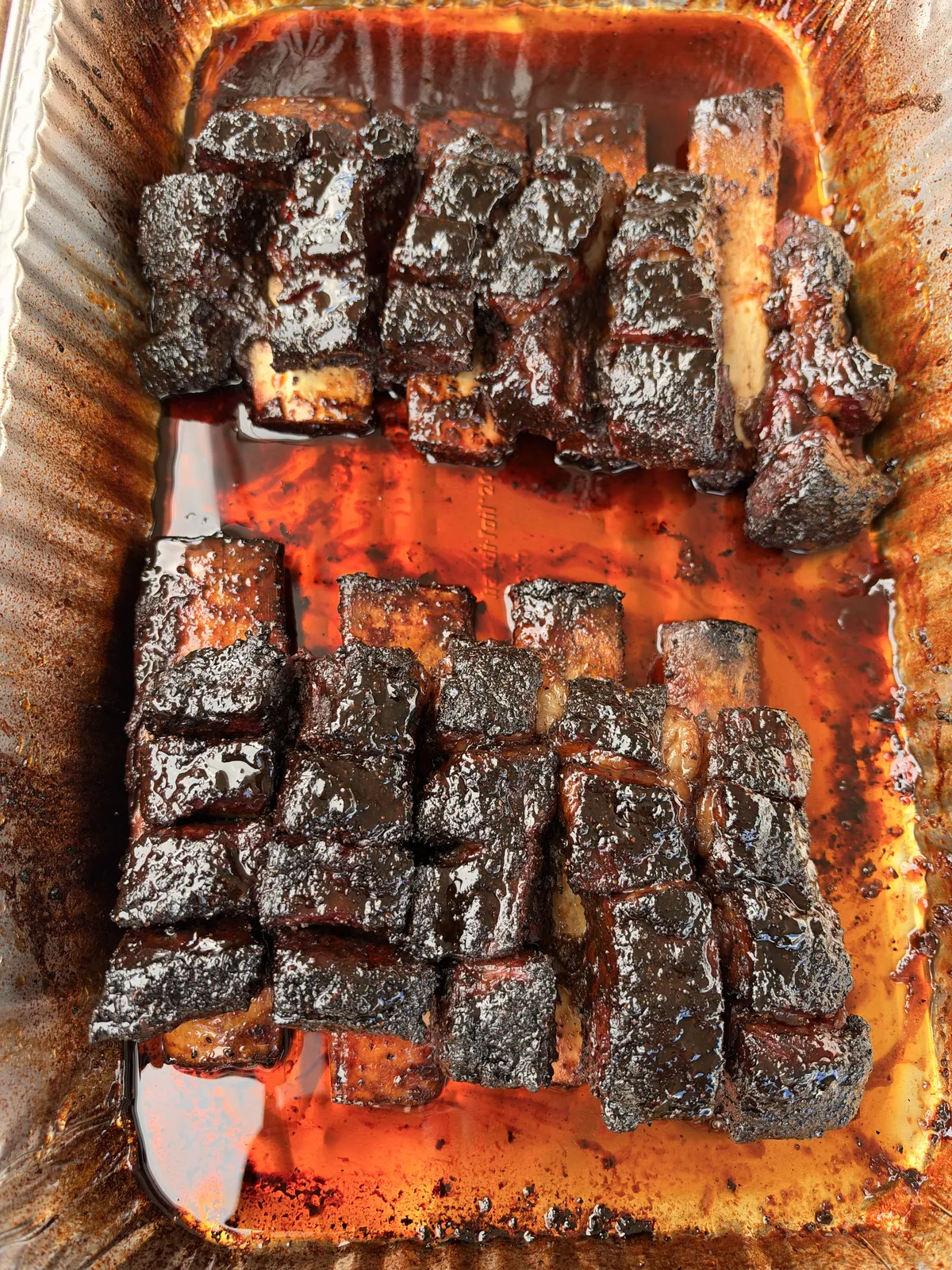 Caramelized BBQ burnt ends coated in dark glossy sauce, arranged in a foil roasting pan with rich juices
