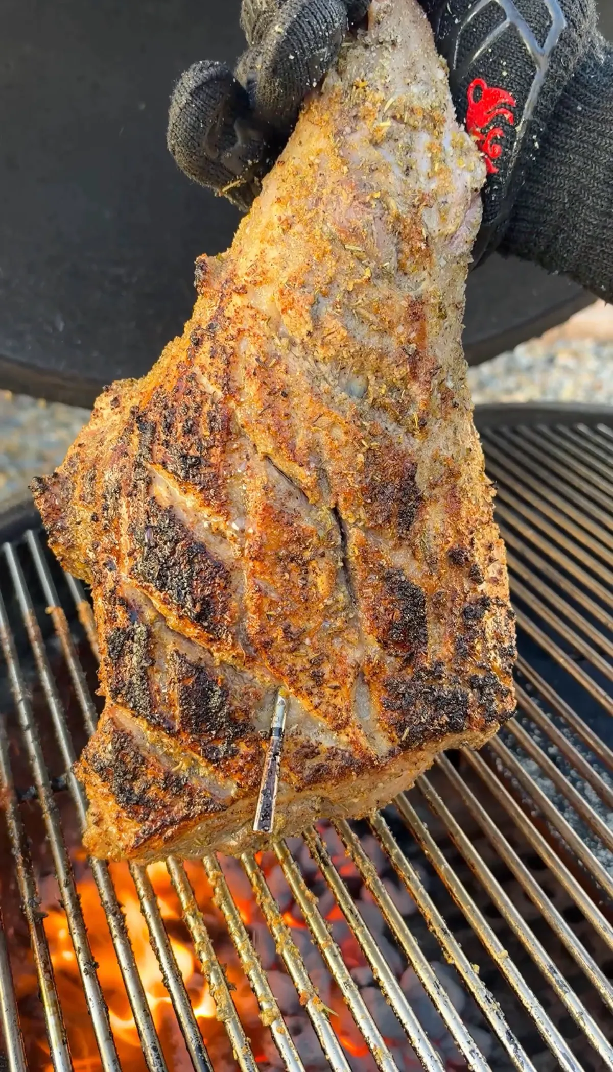 Scored leg of lamb with Greek oregano seasoning crust being seared over hot grill grates with gloved hand