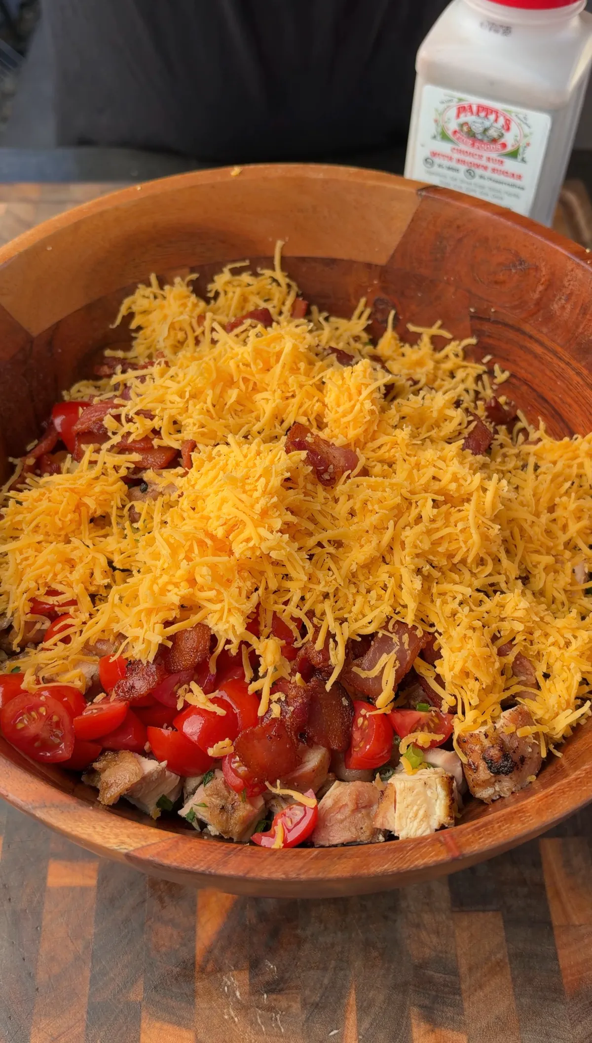 Large wooden bowl with cubed grilled chicken, crispy bacon, cherry tomatoes, and freshly grated cheddar cheese before tossing
