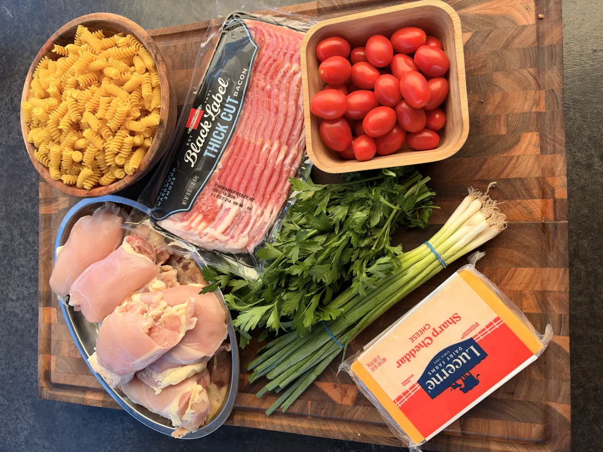 Flat lay of pasta salad ingredients: rotini, thick-cut bacon, cherry tomatoes, chicken thighs, sharp cheddar, green onions, and parsley