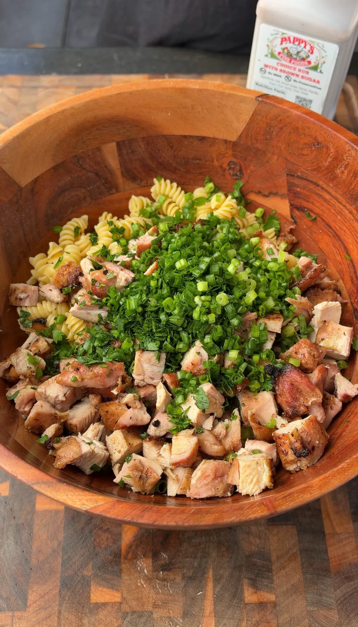 Large wooden bowl with cubed grilled chicken thighs, rotini pasta, chopped green onions, and fresh parsley before tossing