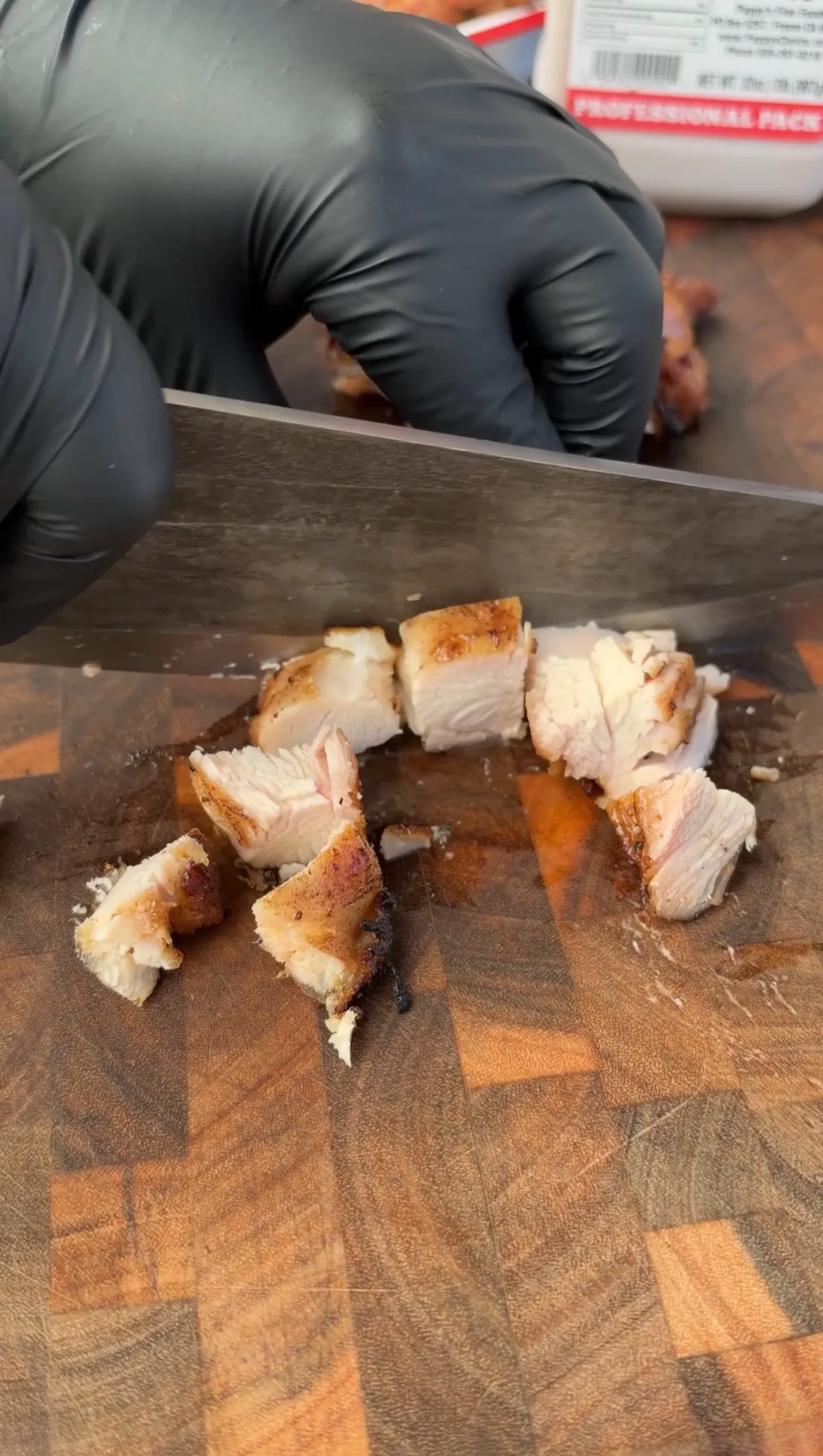 Grilled chicken thighs being cut into bite-sized cubes on a wood cutting board with black gloved hands