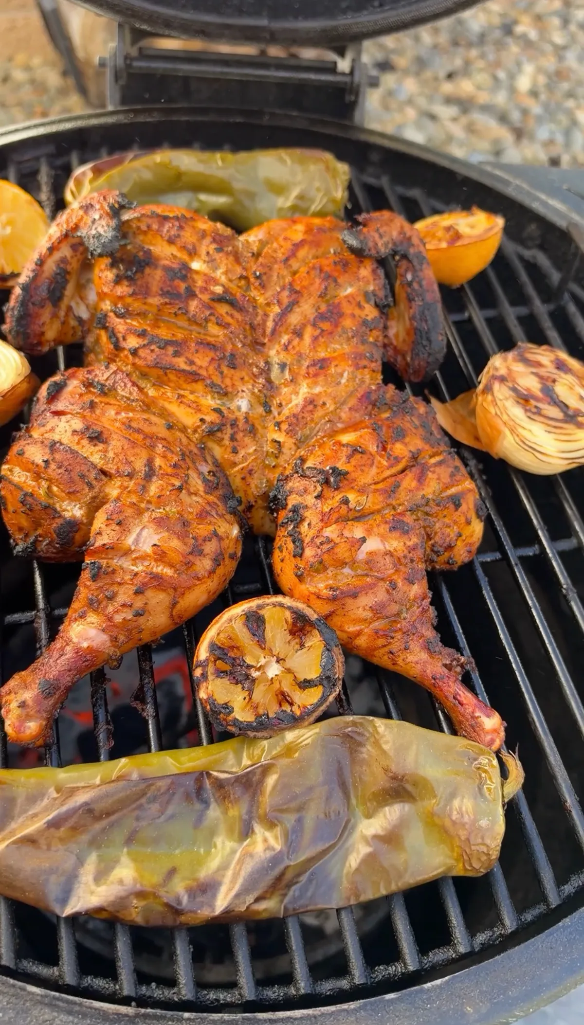 Spatchcocked chicken with chipotle smoked paprika marinade grilling alongside charred onions, lemons, and roasted Anaheim peppers on a charcoal grill