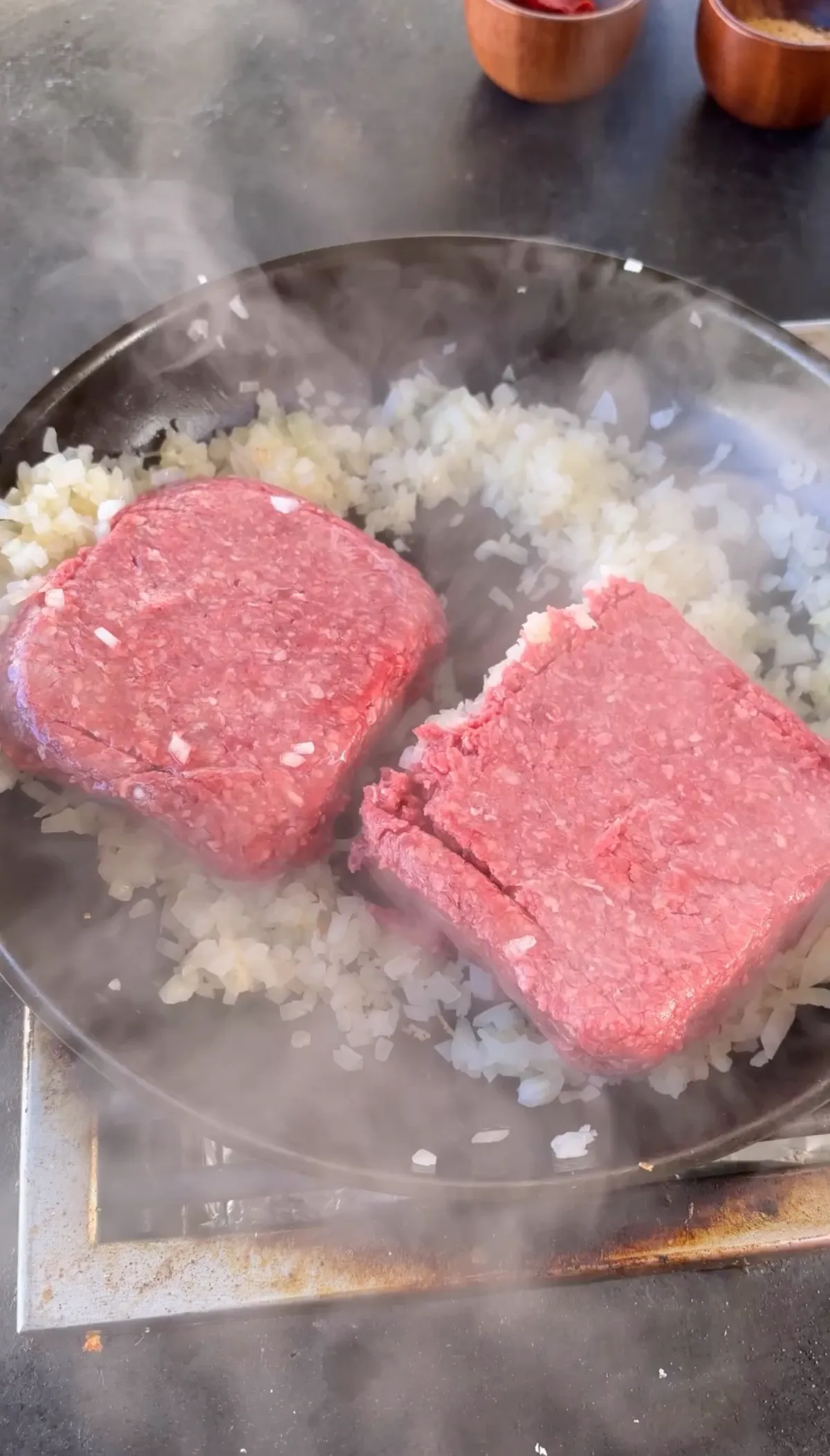 Two 85/15 ground beef patties cooking over sweated diced onions in a steaming pan