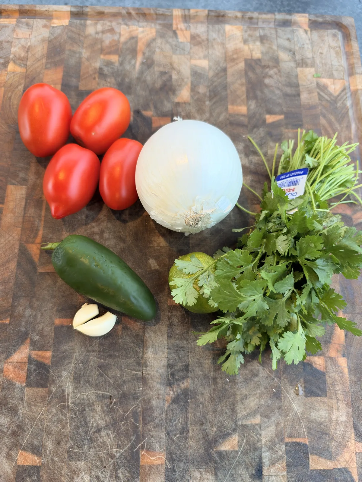 Fresh homemade salsa ingredients: Roma tomatoes, jalapeño, white onion, garlic cloves, lime, and cilantro on a wooden cutting board