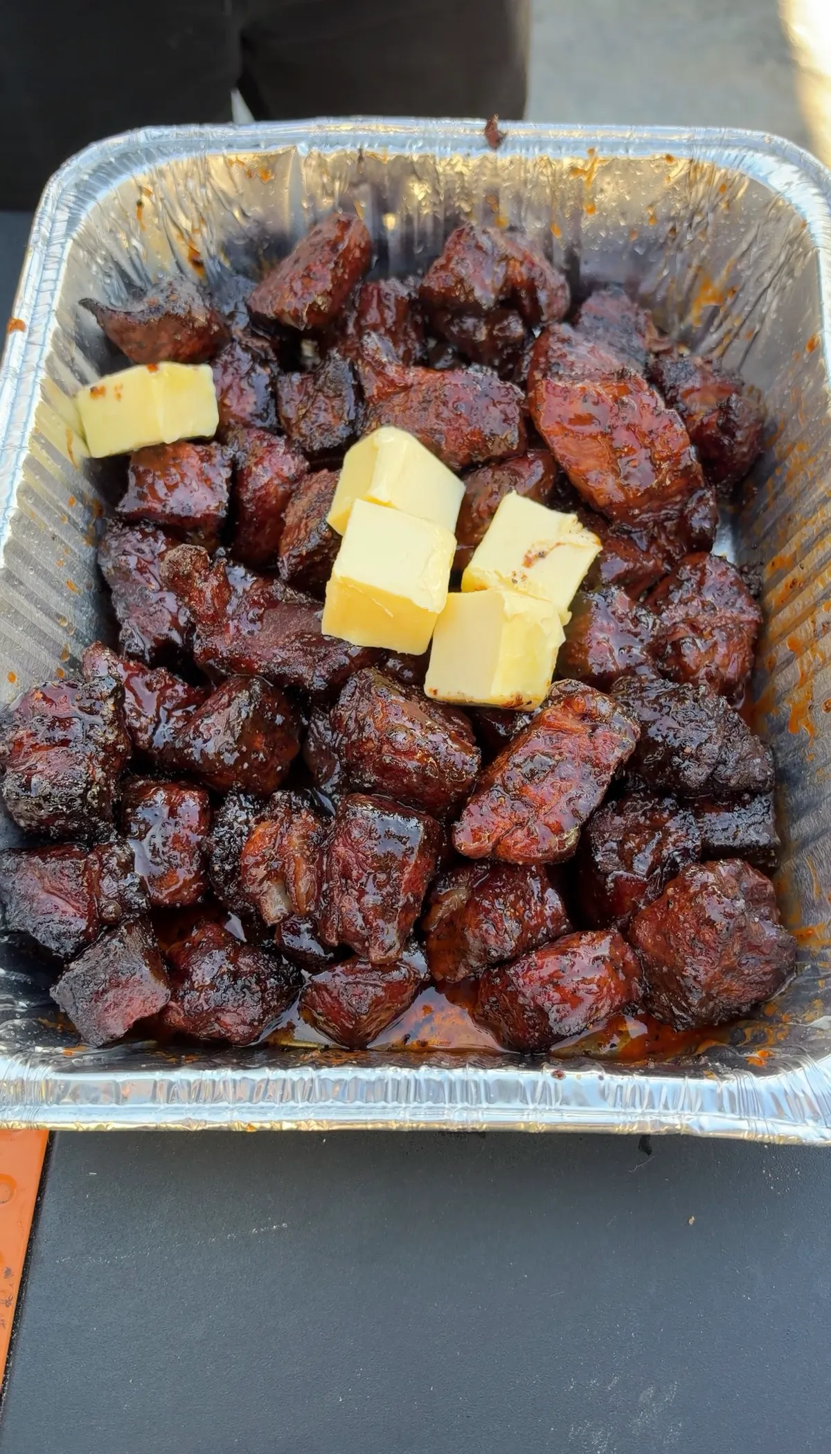 Honey BBQ smoked chuck roast burnt ends glazed in sticky sauce with butter pats in a foil pan