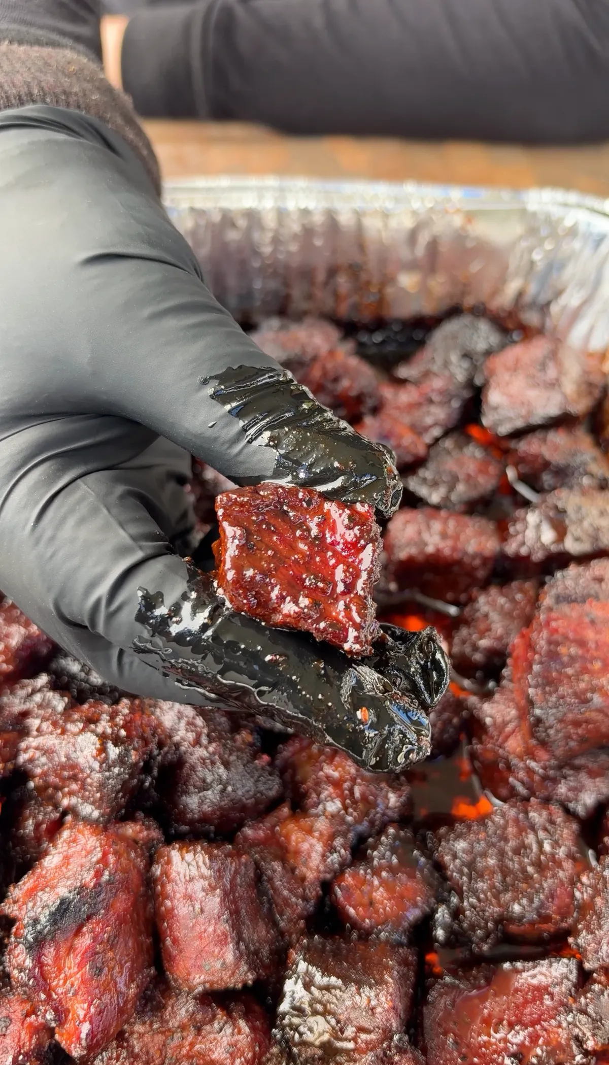 Gloved hand holding a saucy chuck roast burnt end over a foil pan of honey BBQ glazed smoked beef cubes
