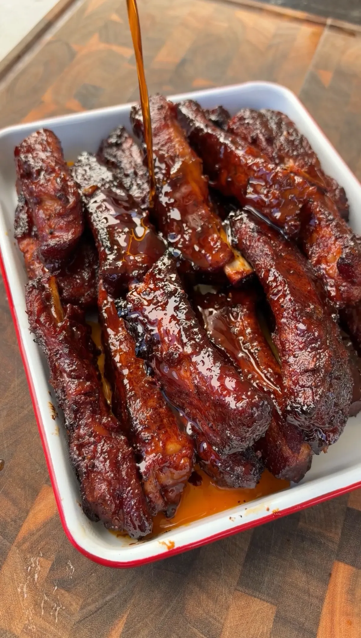 Sticky smoked barbecue party ribs in a white enamel tray with honey-barbecue glaze being drizzled over the top