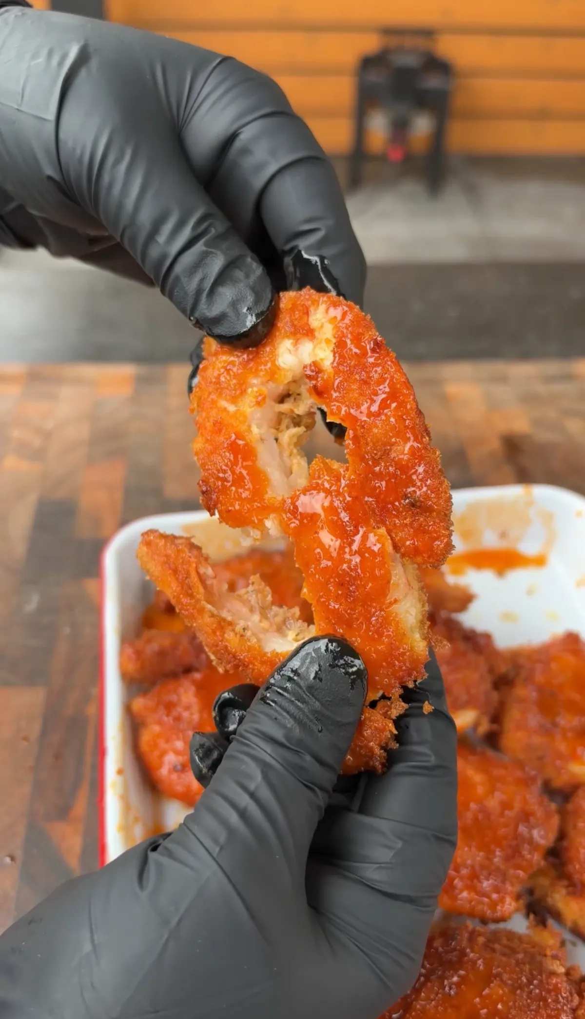 Gloved hands pulling apart panko-breaded shrimp coated in hot honey buffalo sauce over a tray of fried shrimp