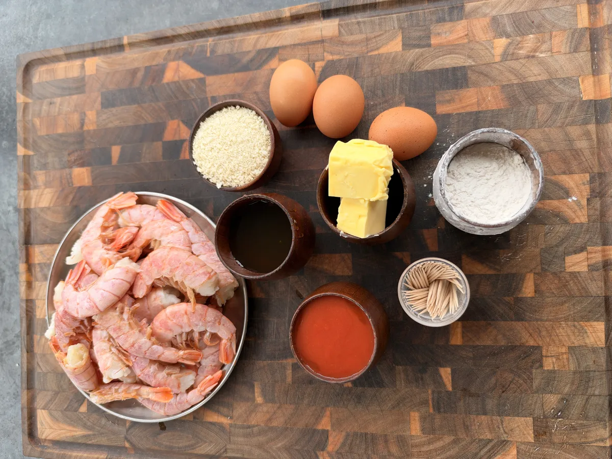 Flat lay of hot honey fried shrimp ingredients: raw shrimp, eggs, butter, panko, flour, buffalo sauce, honey, and toothpicks on a wood board