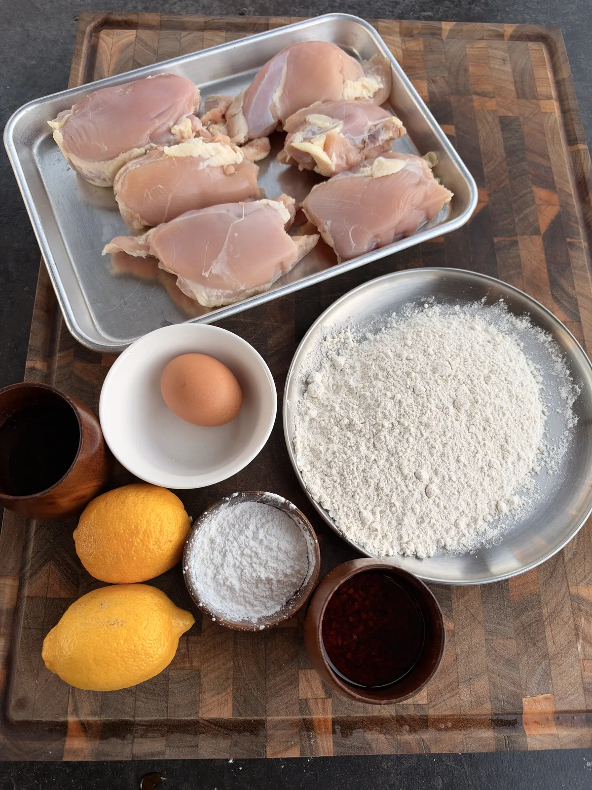 Raw boneless skinless chicken thighs, eggs, all-purpose flour, cornstarch, lemons, hot honey, and chili oil on a wooden cutting board