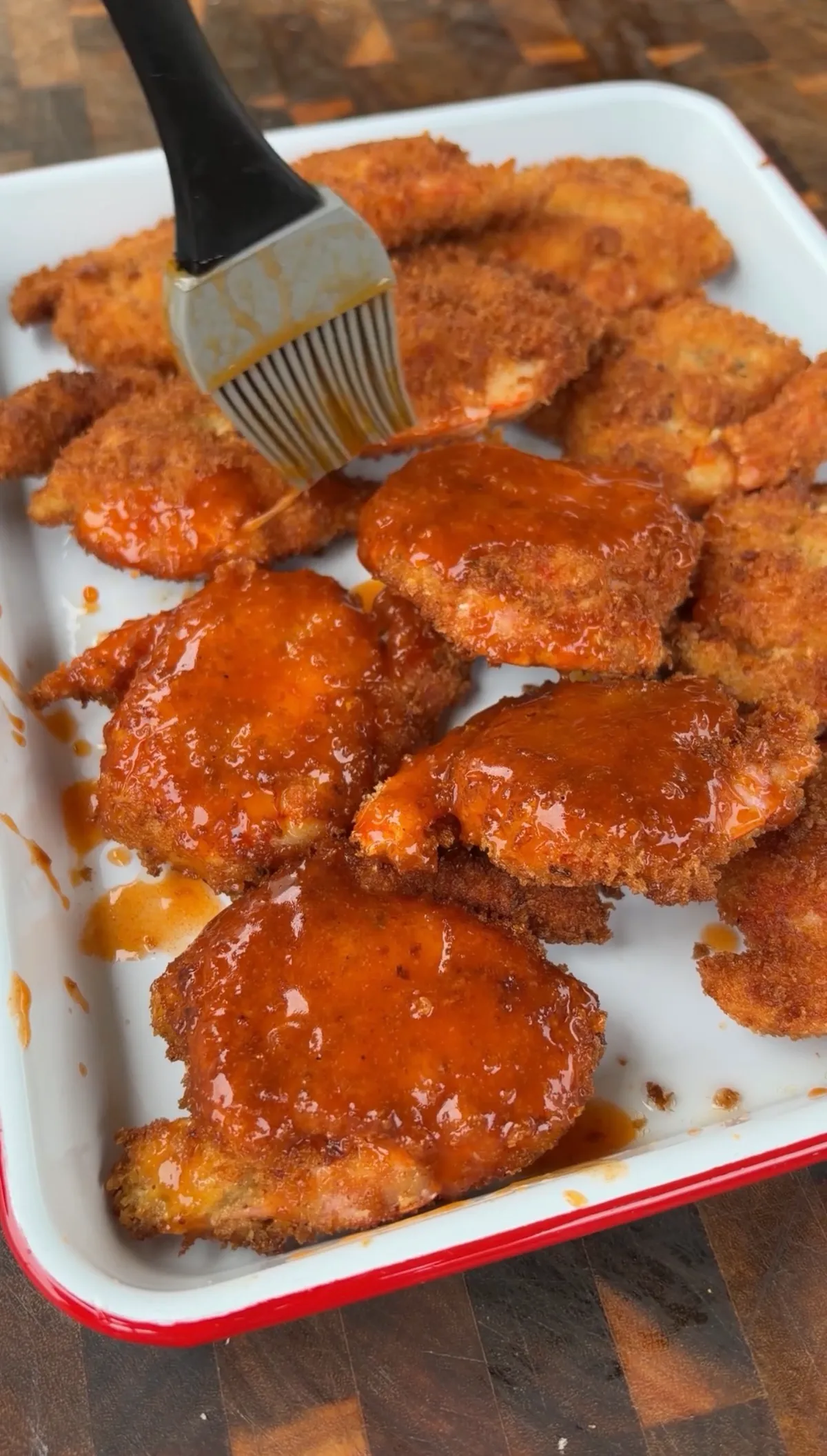 Crispy panko-breaded fried shrimp being brushed with hot honey buffalo sauce on a white enamel tray