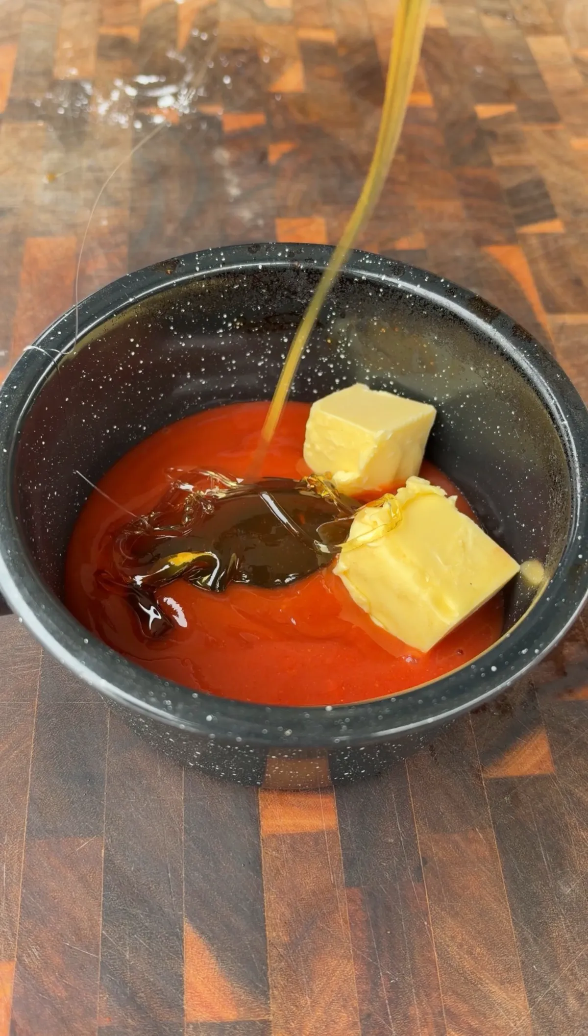 Unsalted butter pats and honey being added to buffalo wing sauce in a dark saucepan for hot honey sauce