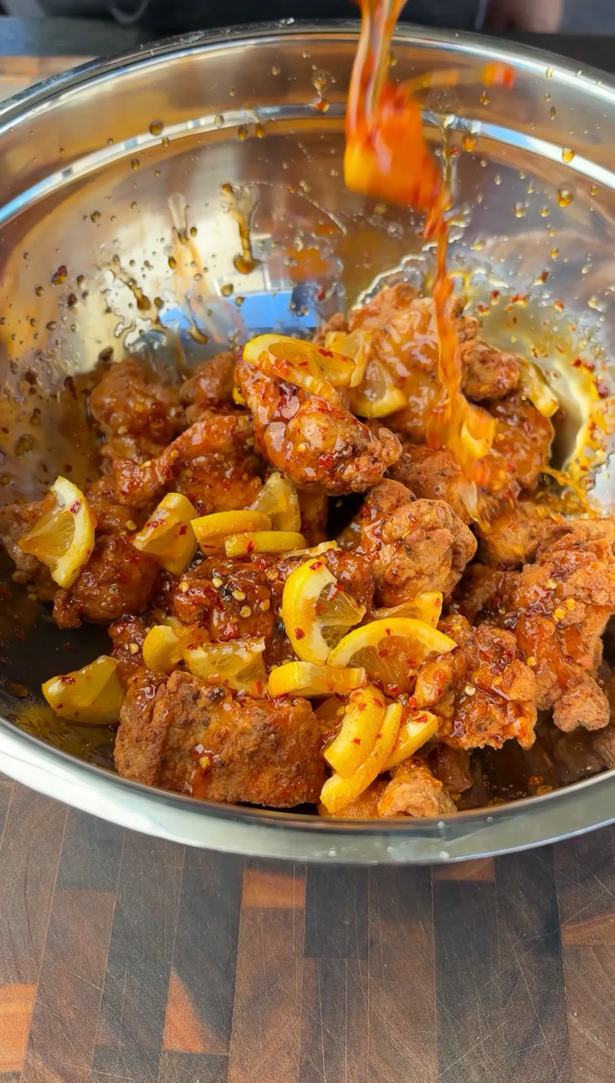 Hot honey lemon pepper sauce being poured over crispy fried chicken thigh nuggets with lemon quarters in a stainless steel mixing bowl