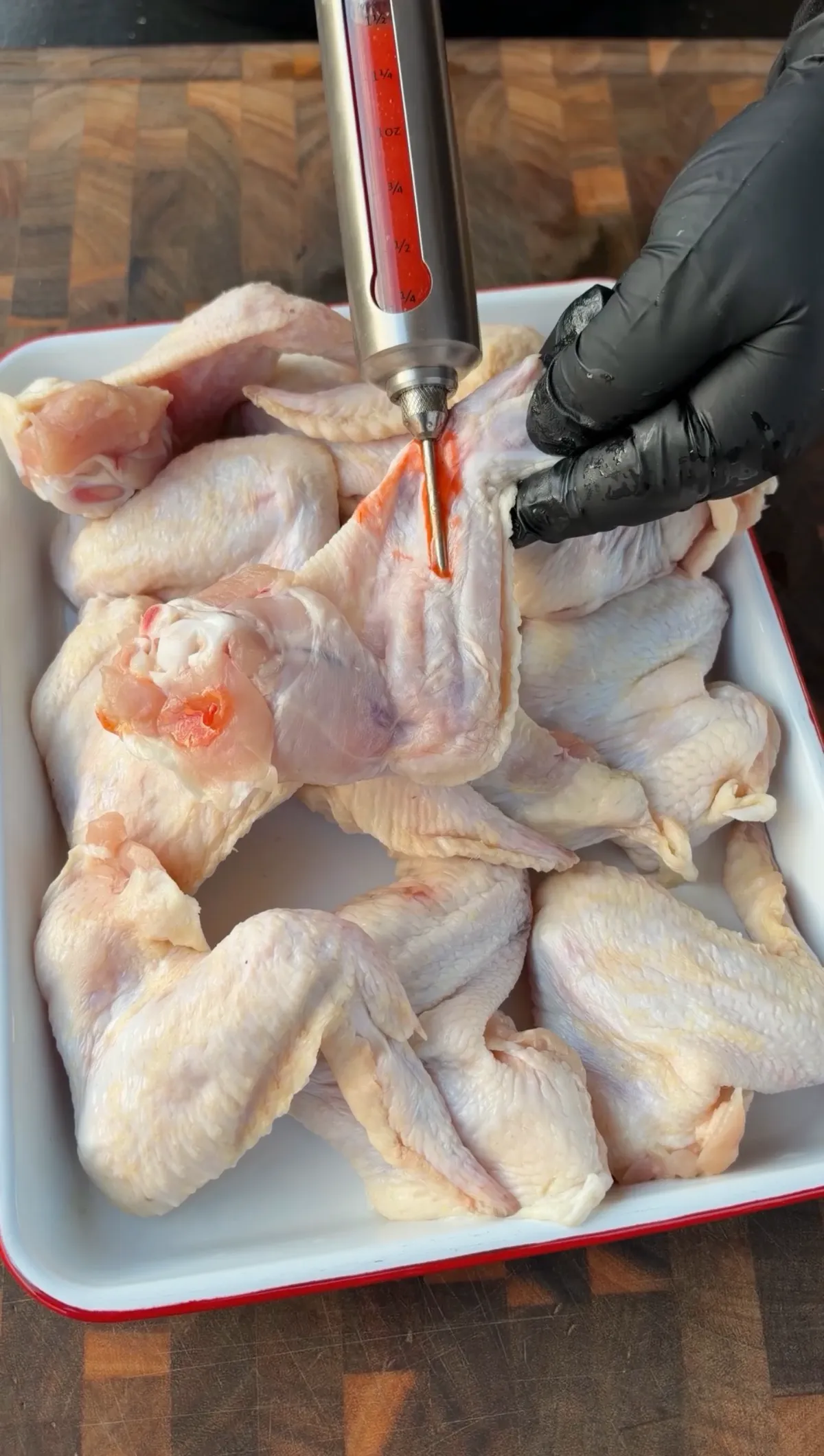 Hands in black gloves injecting jumbo chicken wings with red wing sauce and butter mixture in a white pan