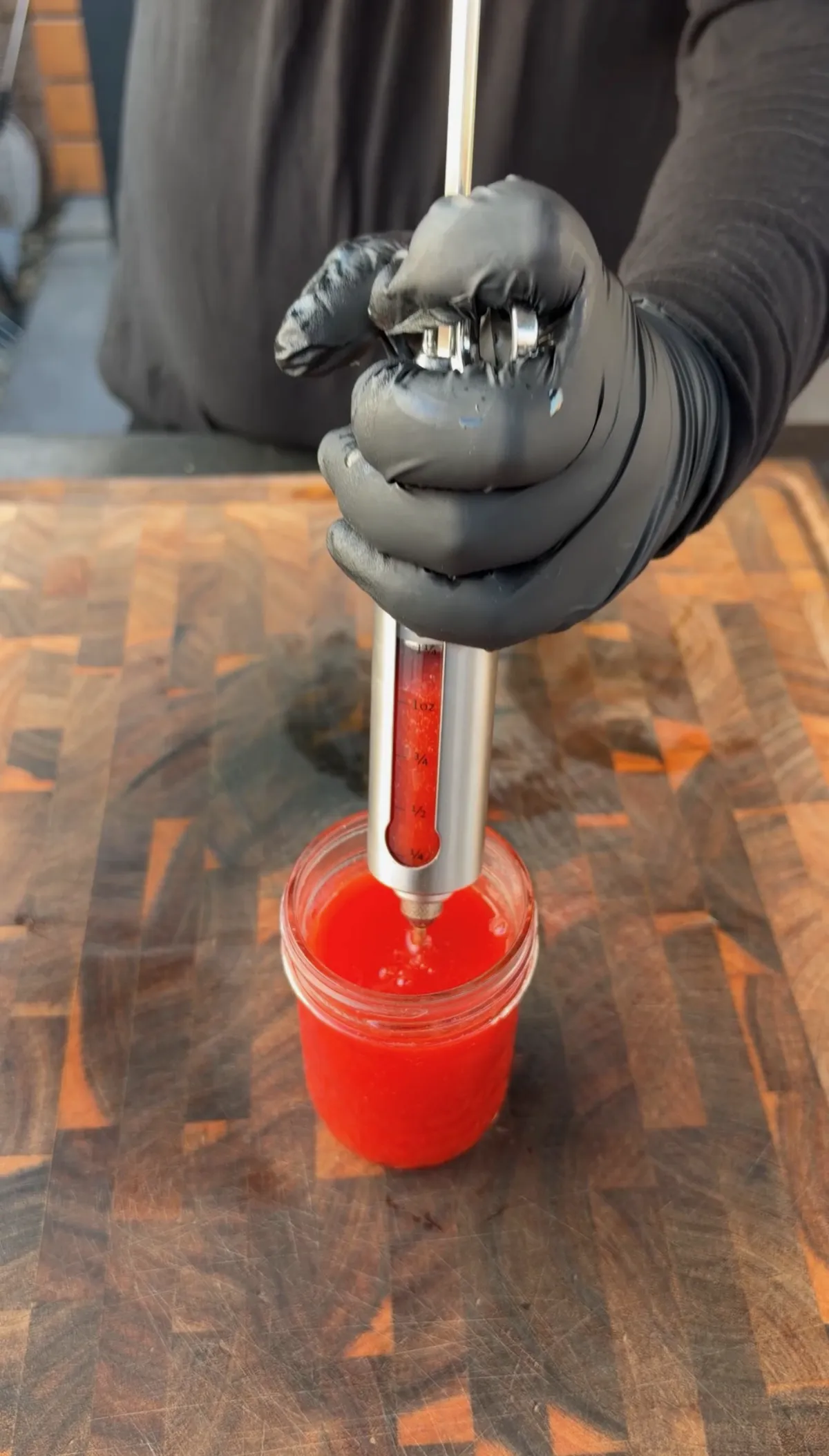 Gloved hands using a meat injector to inject red wing sauce mixture into chicken wings in a glass jar