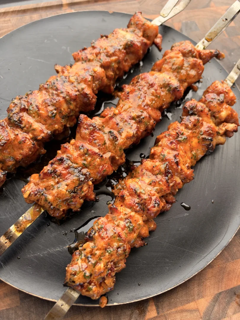 Four grilled chicken thigh skewers coated in glistening cowboy butter with parsley on a dark round platter