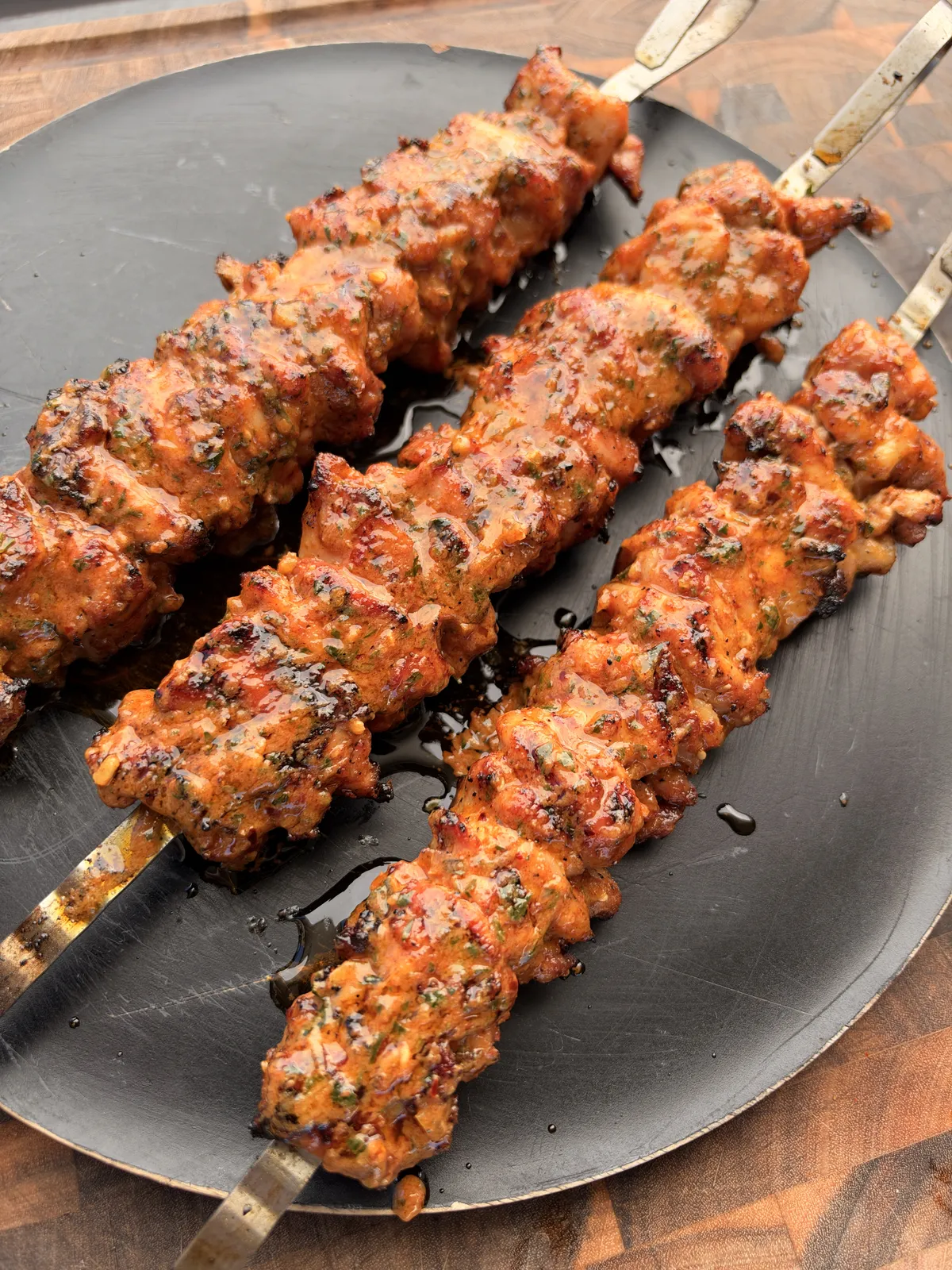 Four grilled chicken thigh skewers coated in glistening cowboy butter with parsley on a dark round platter