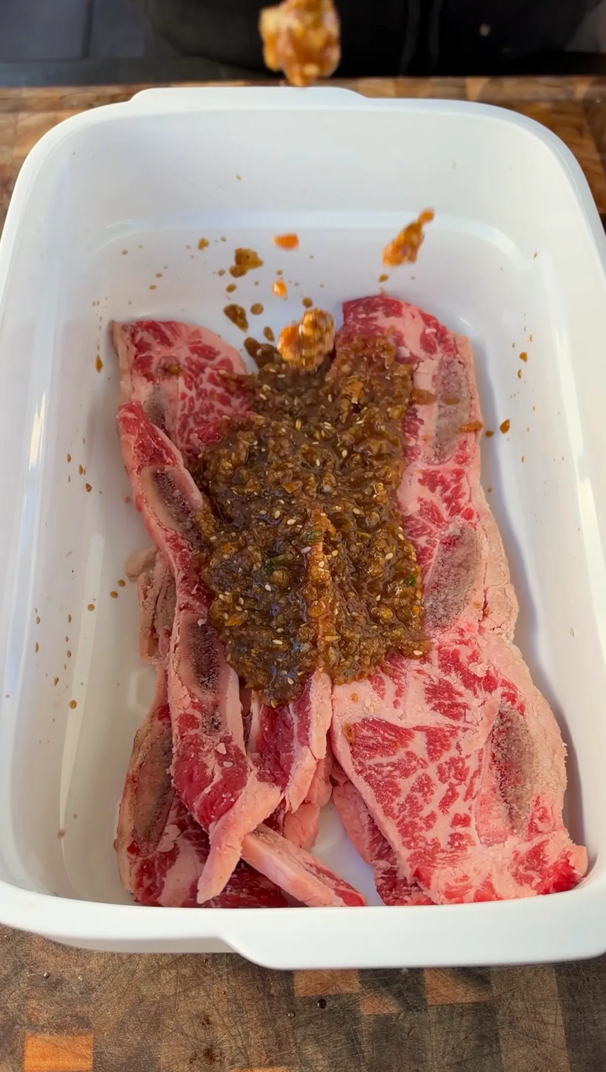 Kalbi marinade with sesame seeds poured over raw flanken-cut beef short ribs in a white container