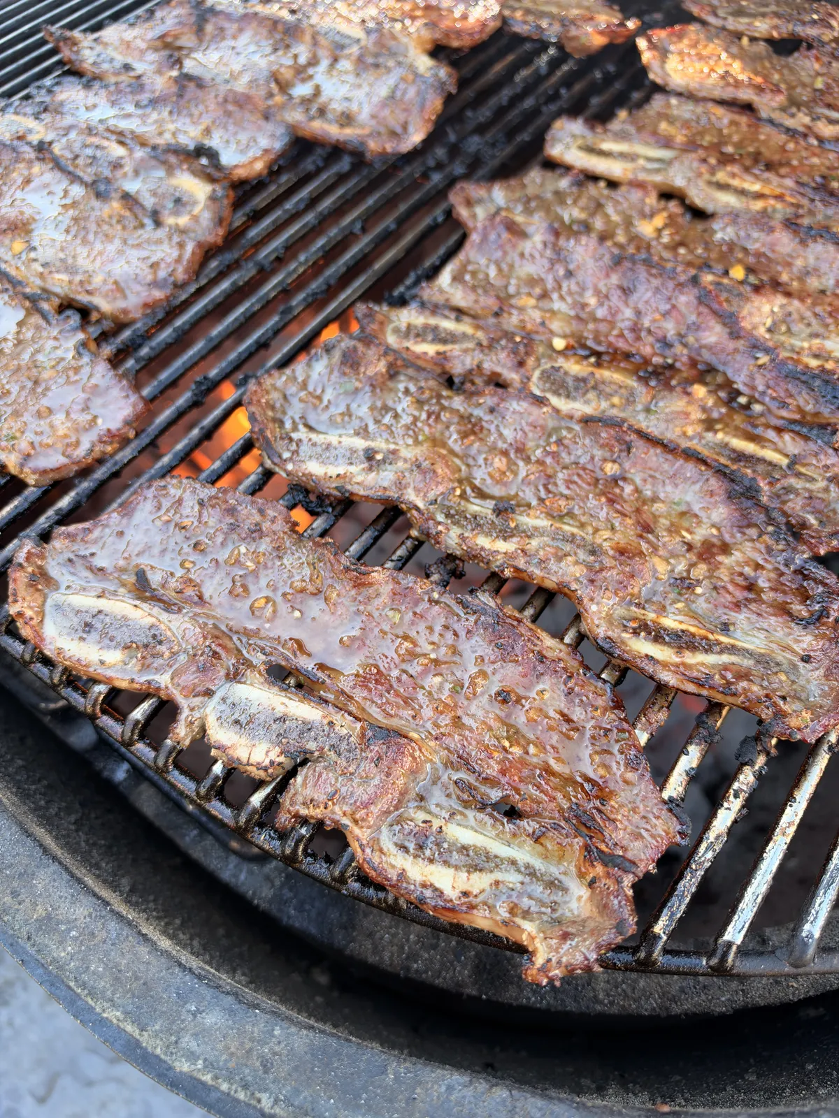 Flanken-cut Korean kalbi beef short ribs grilling over direct high heat on a charcoal grill, showing caramelized soy and sesame marinade glaze