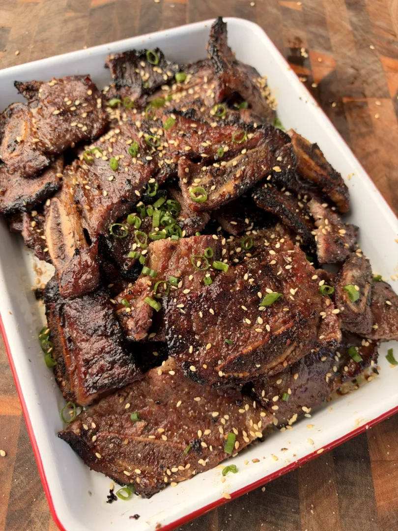 Korean Kalbi beef short ribs piled in a white enamel tray, garnished with toasted sesame seeds and sliced green onions
