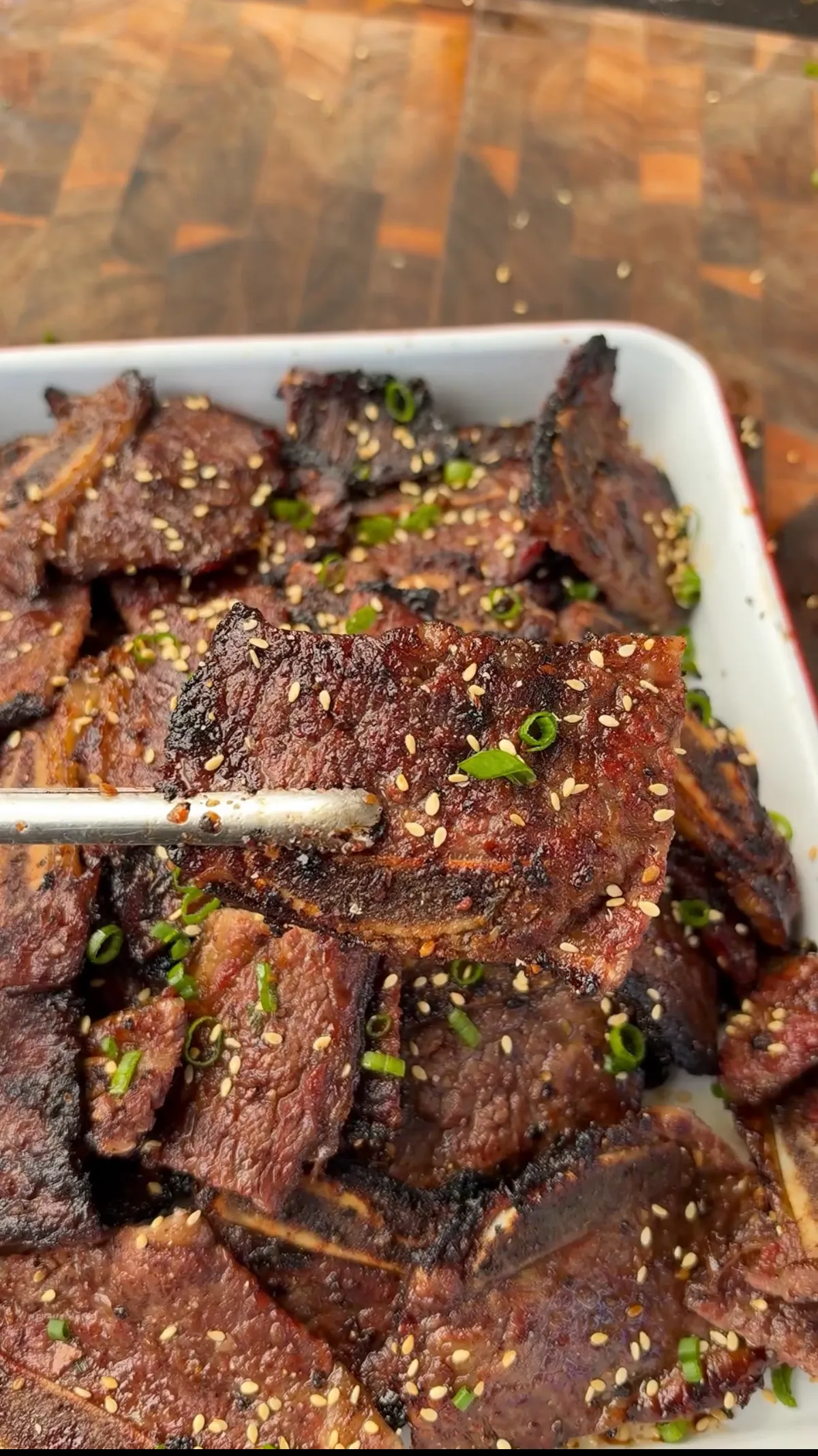 Tongs holding a piece of charred Korean Kalbi beef rib over a white dish of grilled flanken ribs topped with toasted sesame seeds and sliced green onions