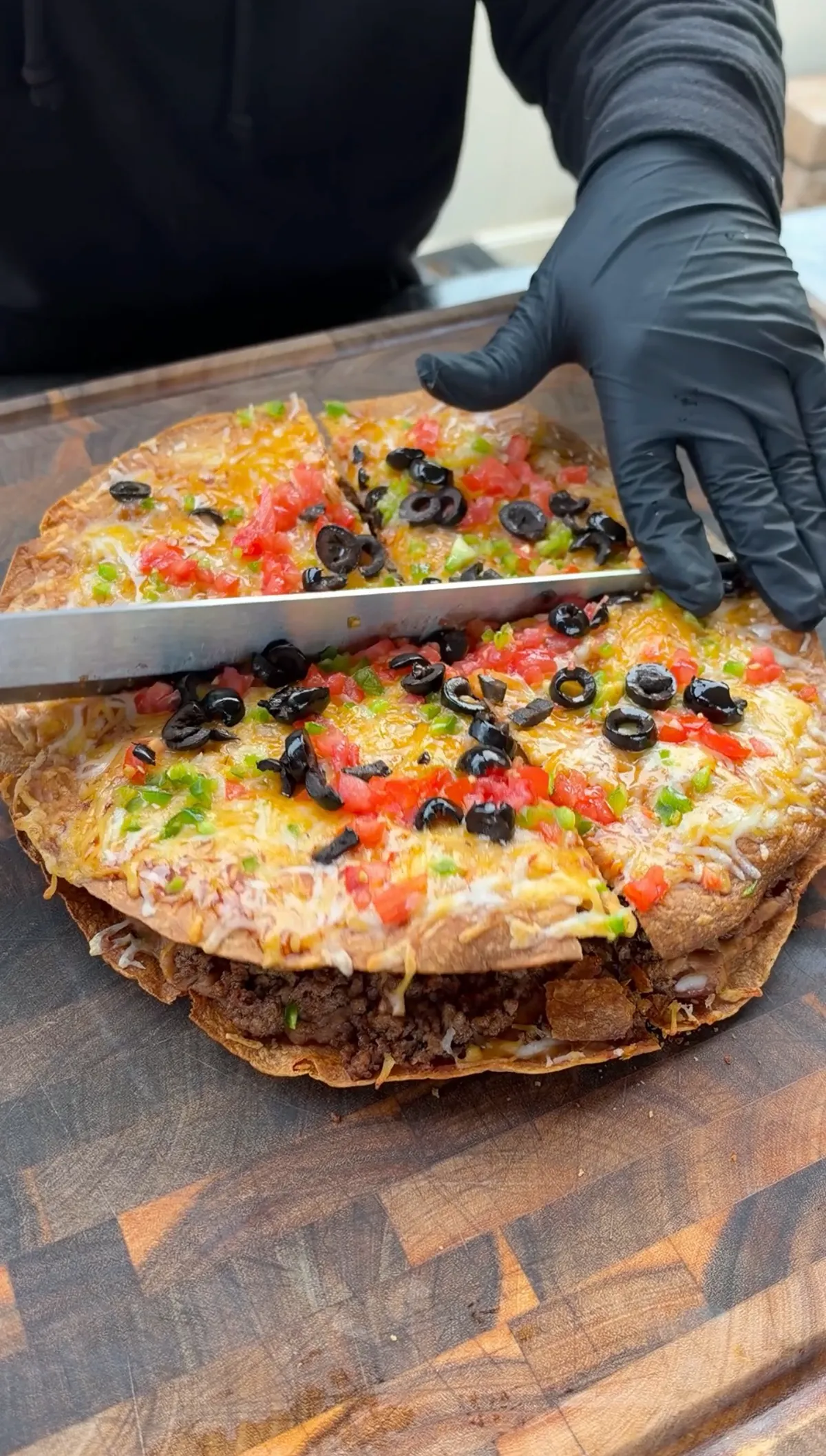 Gloved hands using a knife to slice a layered Mexican pizza topped with Colby Jack cheese, diced tomatoes, black olives and jalapeños