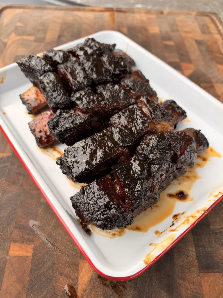 Glossy, caramelized beef short rib burnt ends piled on a white enamel tray with red rim on a wood cutting board