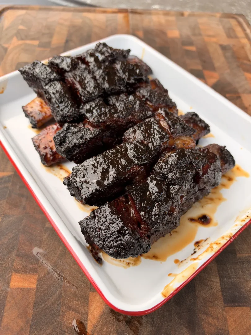 Glossy, caramelized beef short rib burnt ends piled on a white enamel tray with red rim on a wood cutting board